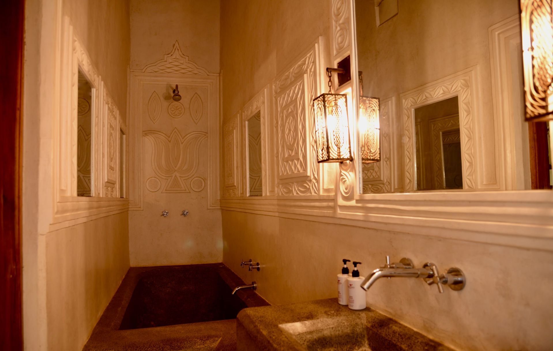 A narrow, cream-colored bathroom with a sink, mirror, wall sconces, and a bathtub.