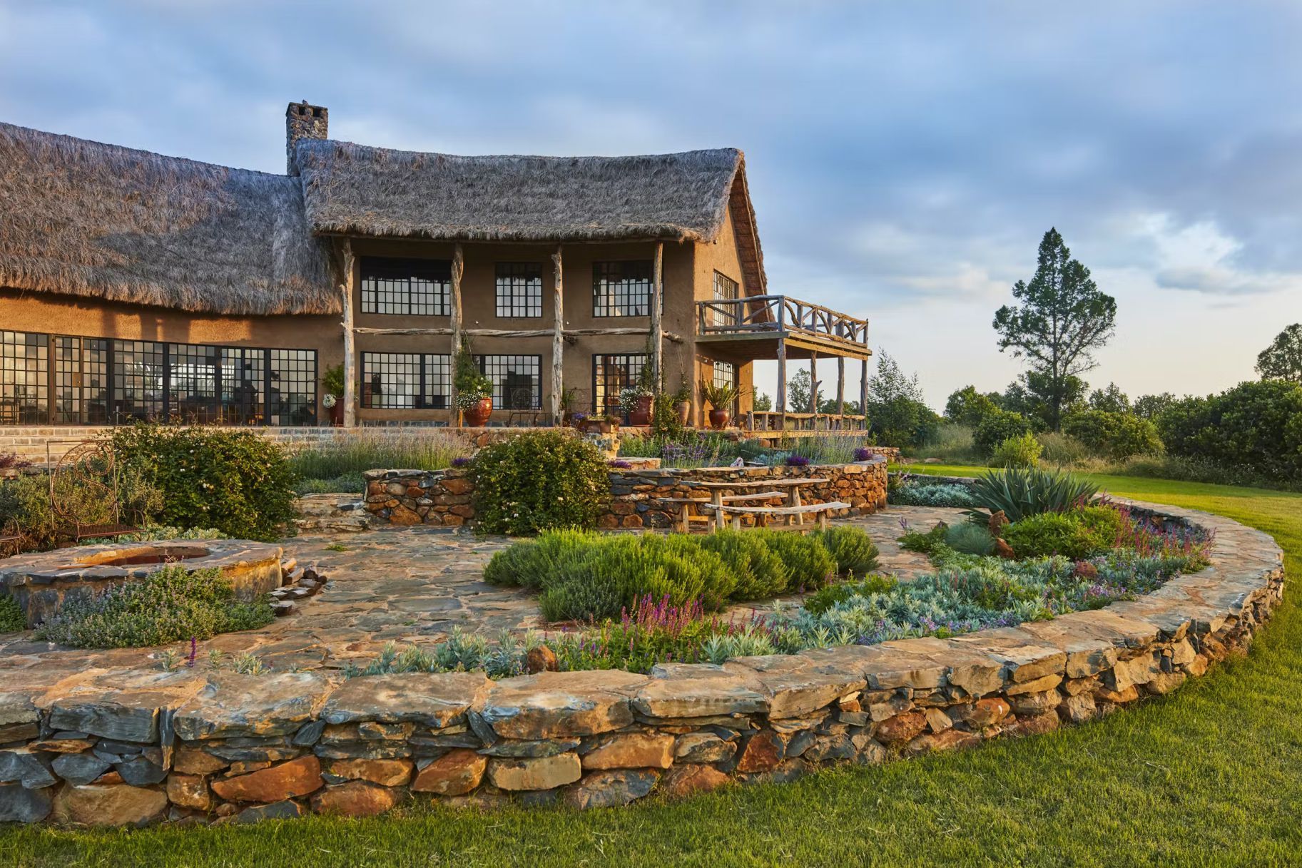 Rustic stone house with thatched roof and landscaped garden under a cloudy sky