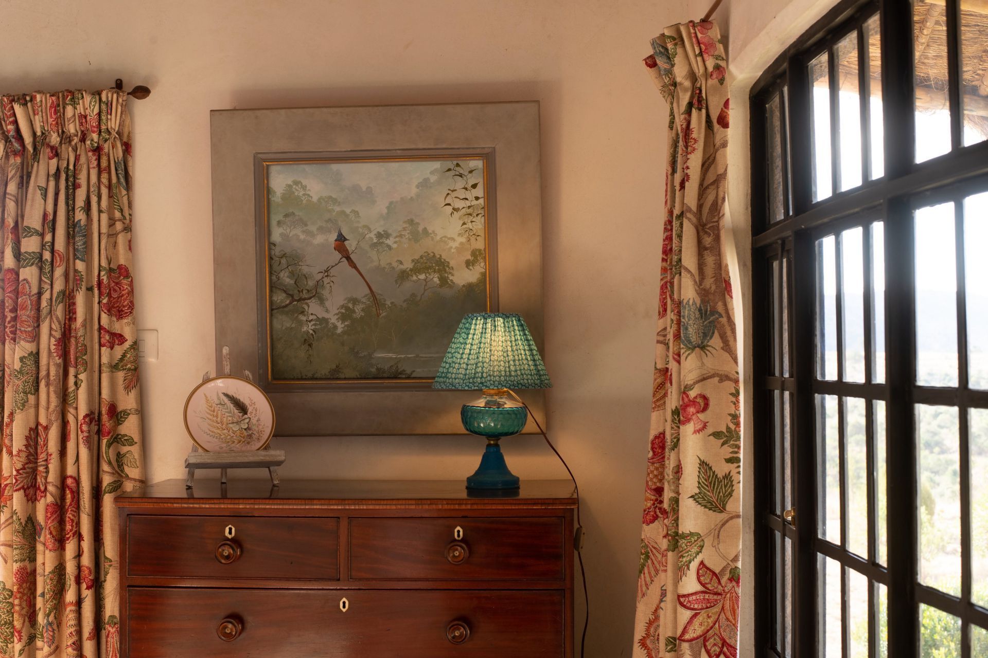 Wooden dresser with lamp and framed painting beside a sunlit window and floral curtains