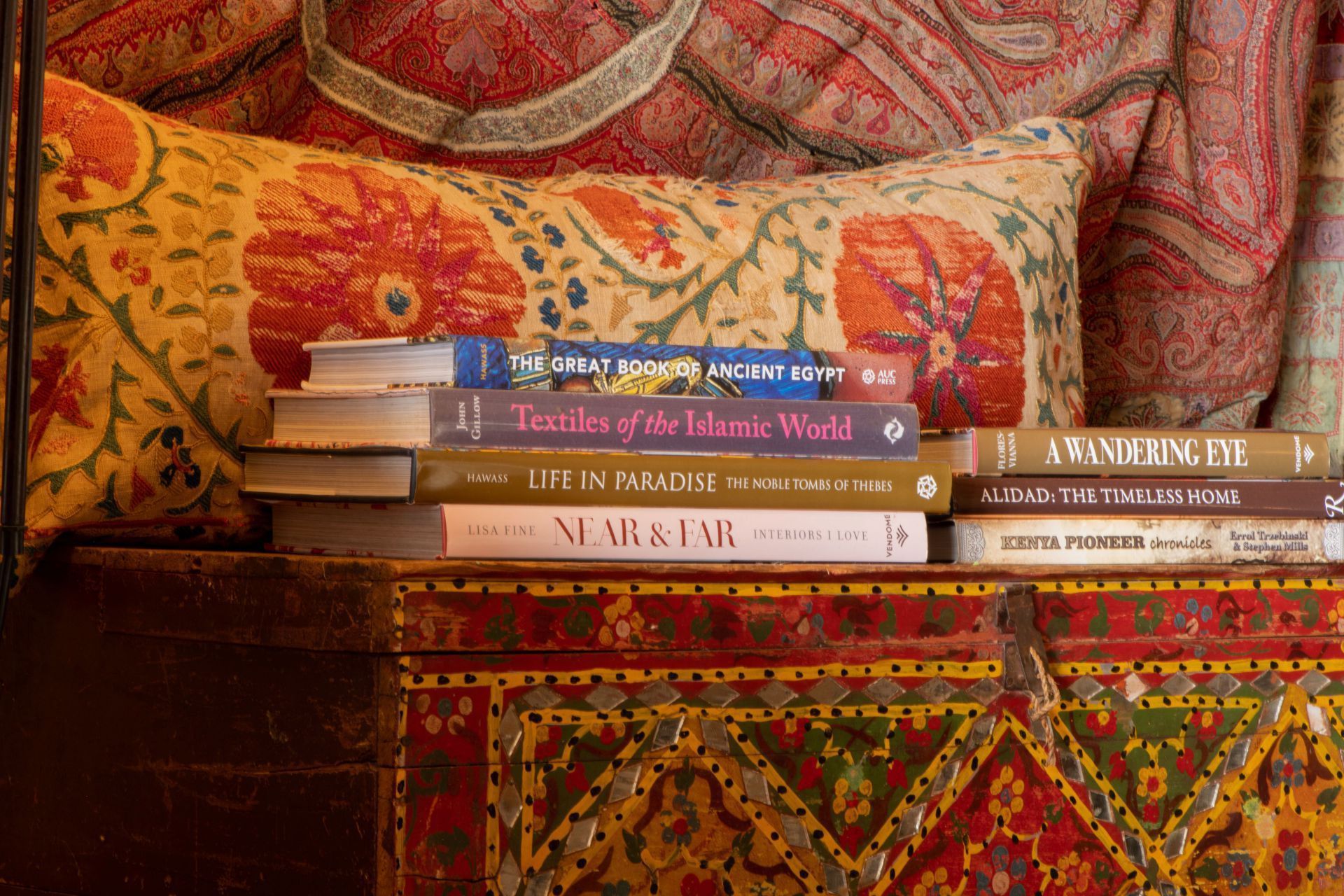 Stack of colorful books on a patterned couch with a floral pillow and red-orange textile backdrop