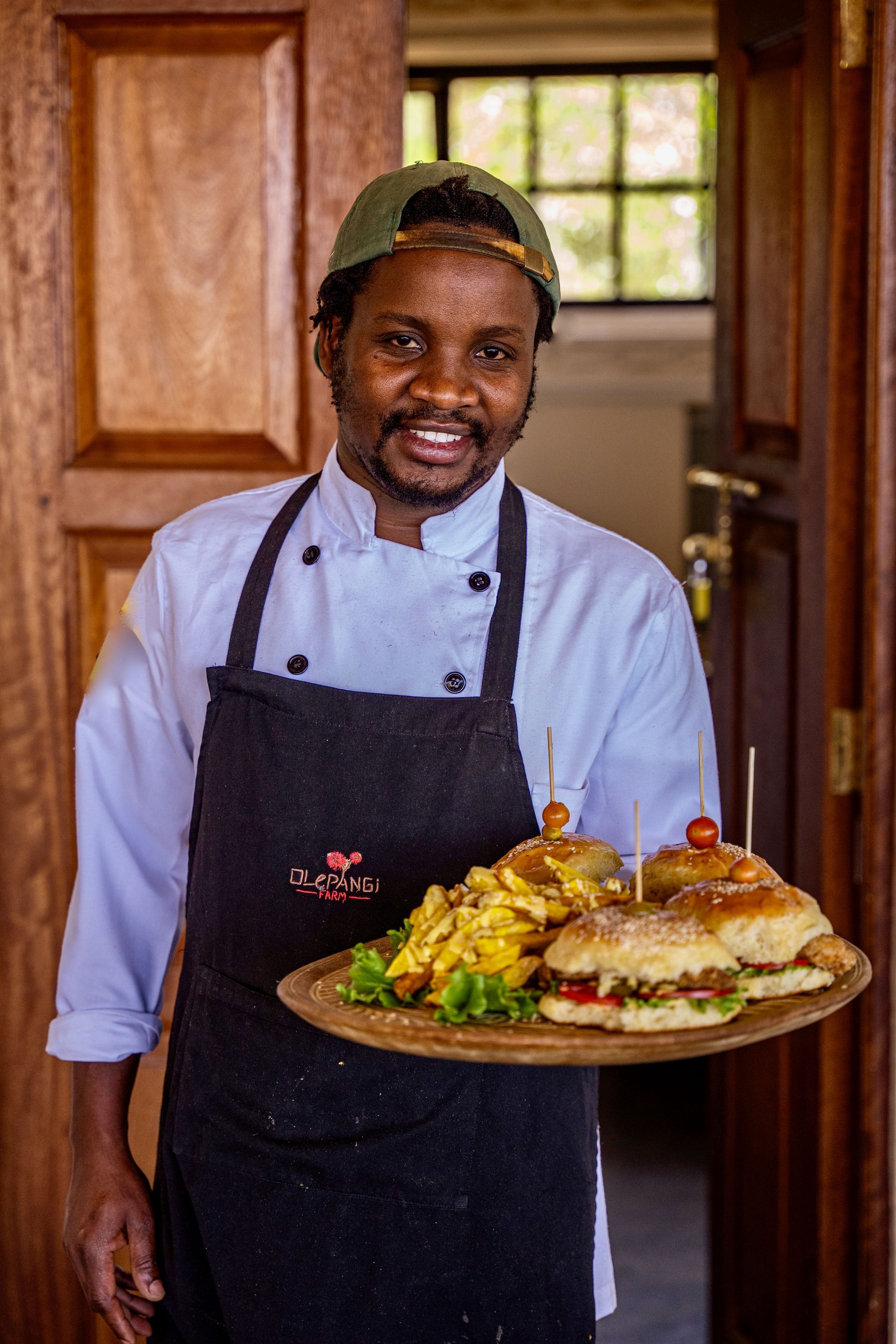 Chef in a kitchen doorway holding a platter of plated food and smiling