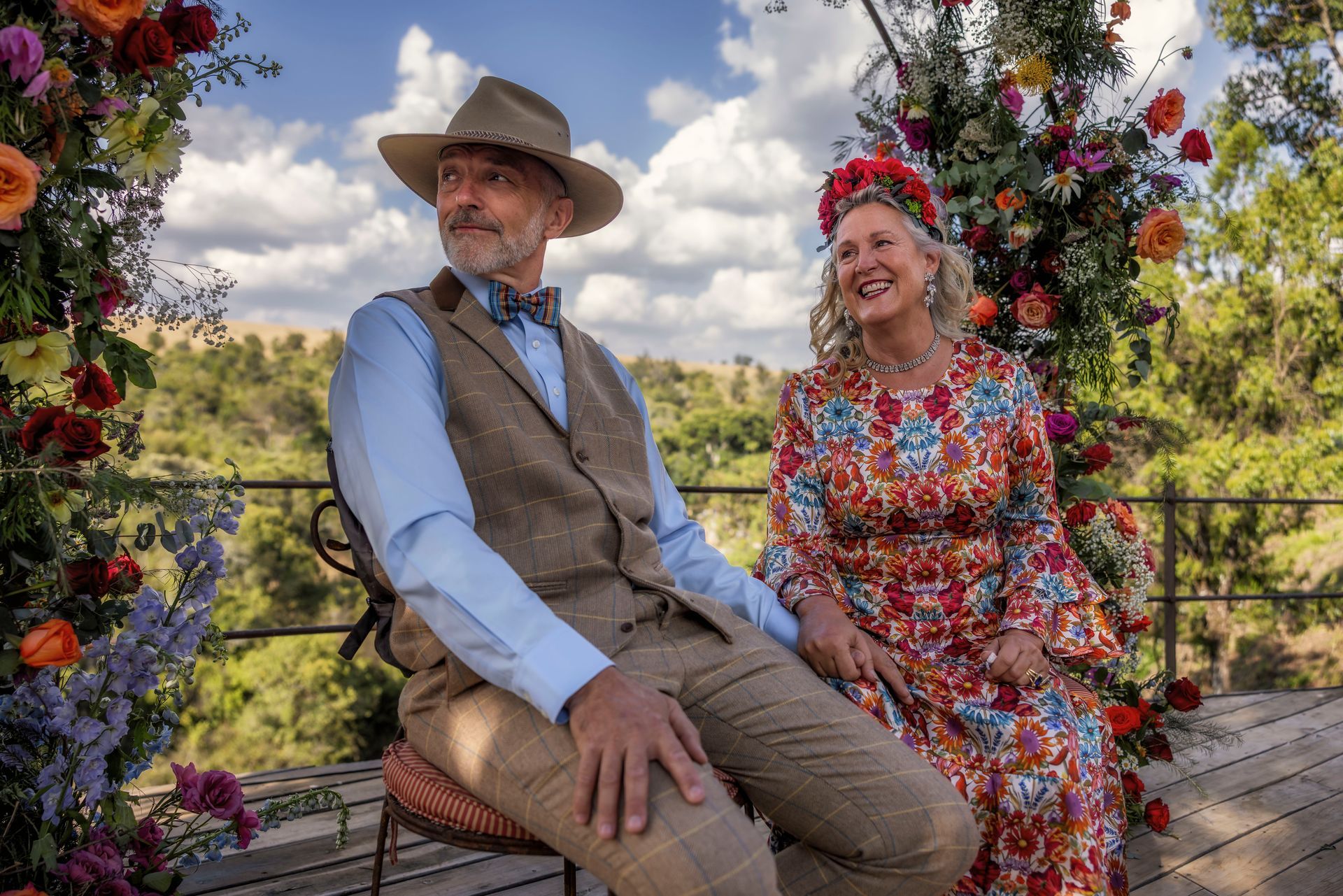Two people in colorful outfits sit on a bench outdoors among flowers and a cloudy sky.