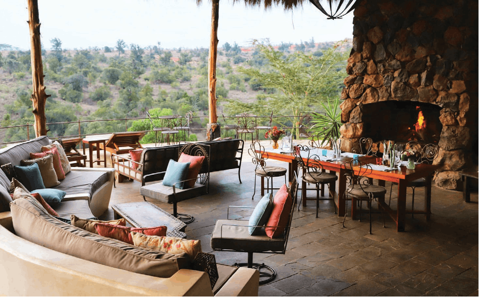 Open-air safari lodge lounge with stone fireplace, wicker chairs, and valley view