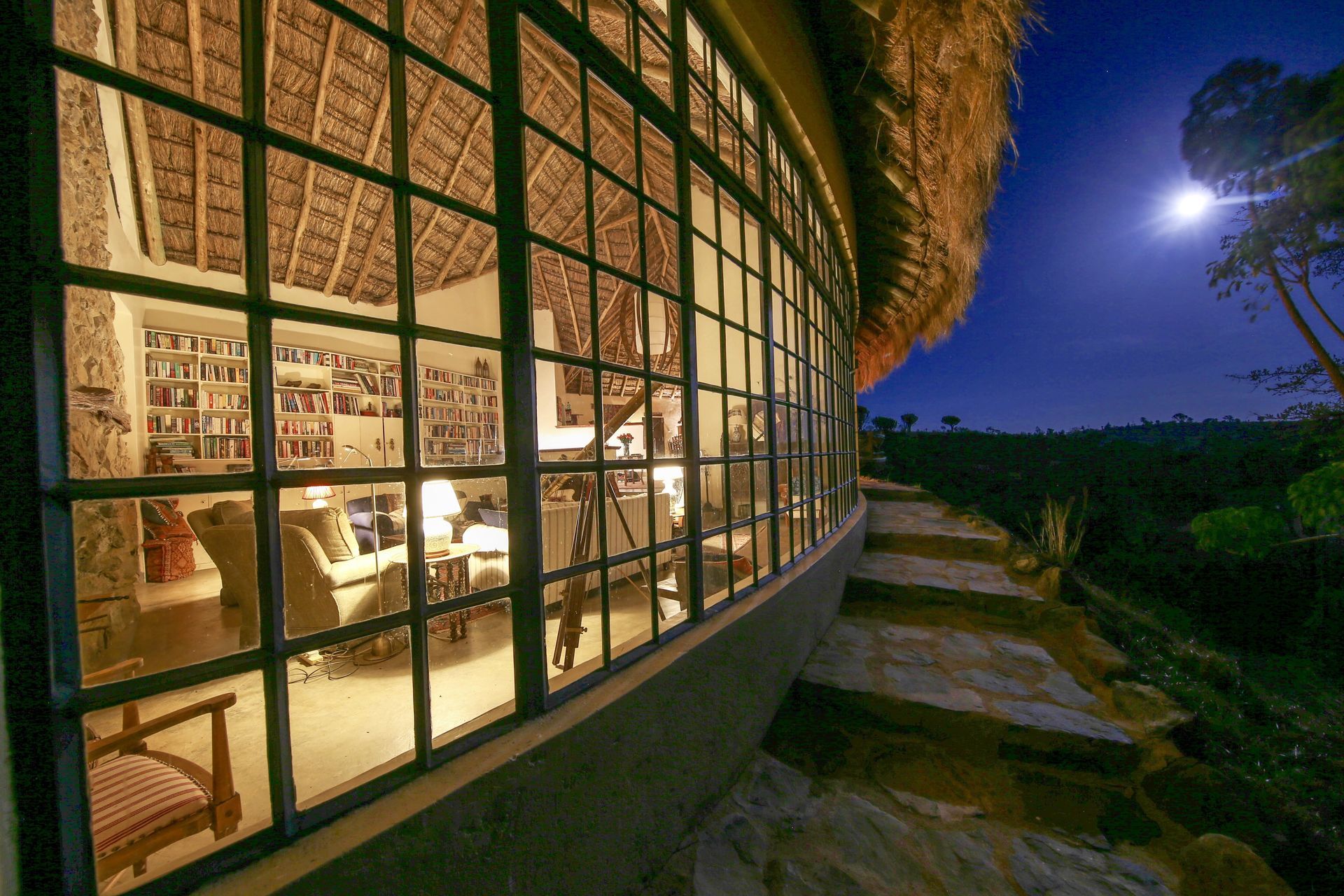 Curved glass-walled building glowing at night, with warm interior lights and a moonlit outdoor path.