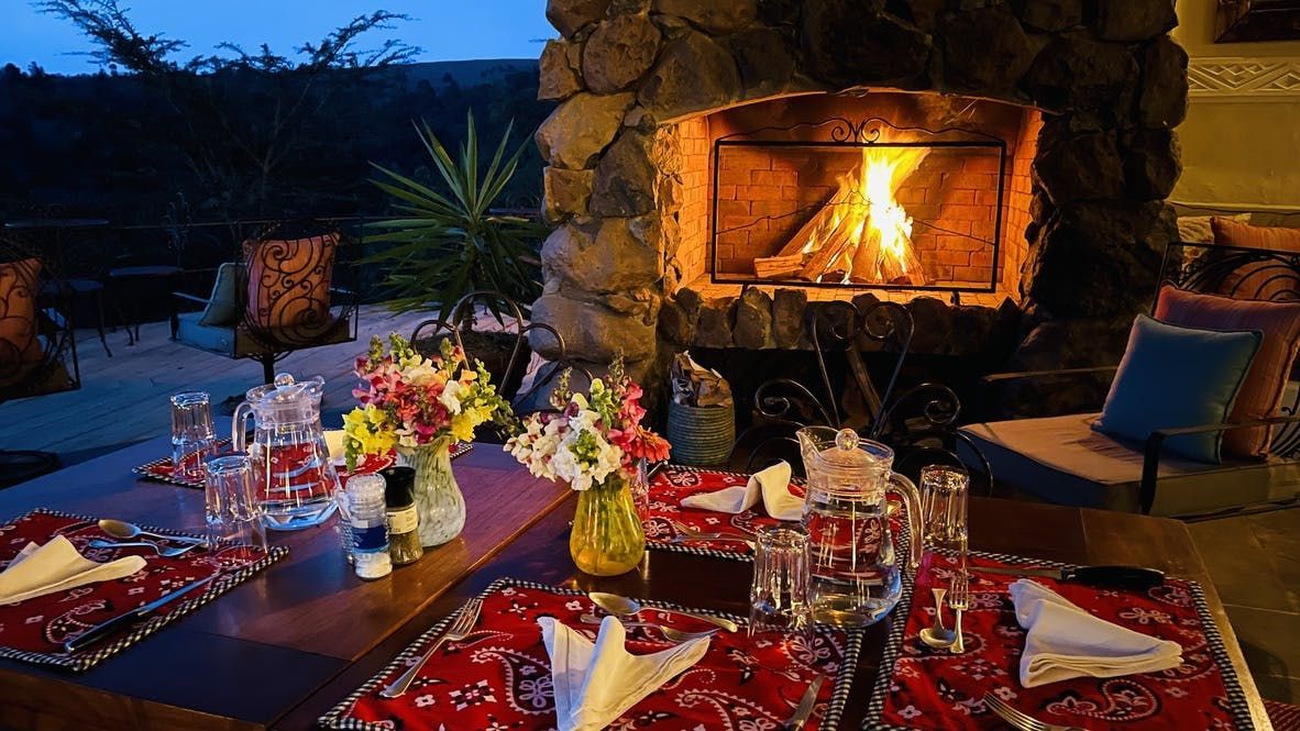 Elegant outdoor dining table with red napkins, flowers, and a lit stone fireplace at dusk