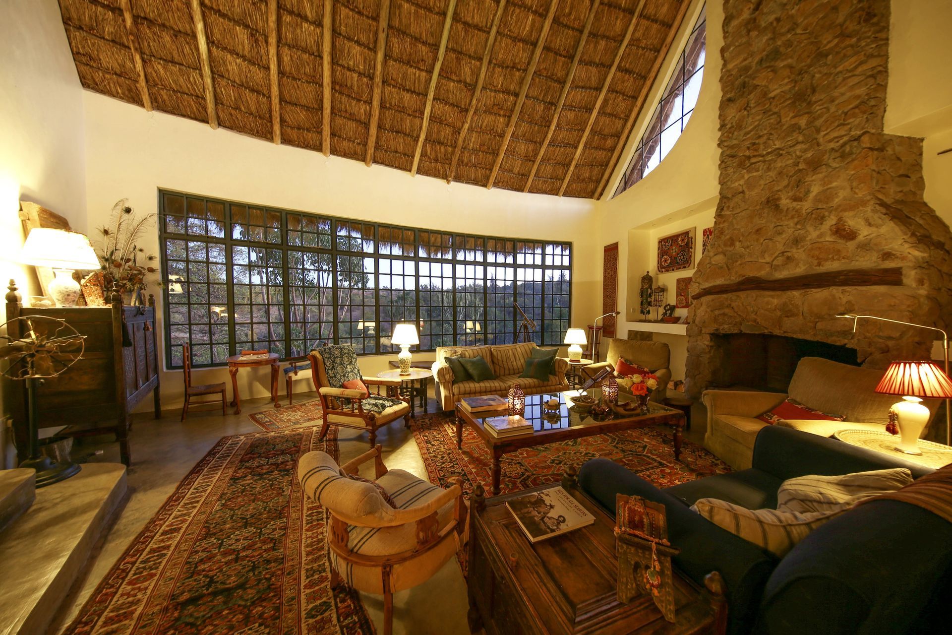 Cozy lodge living room with woven ceiling, stone fireplace, patterned rugs, and large windows overlooking trees