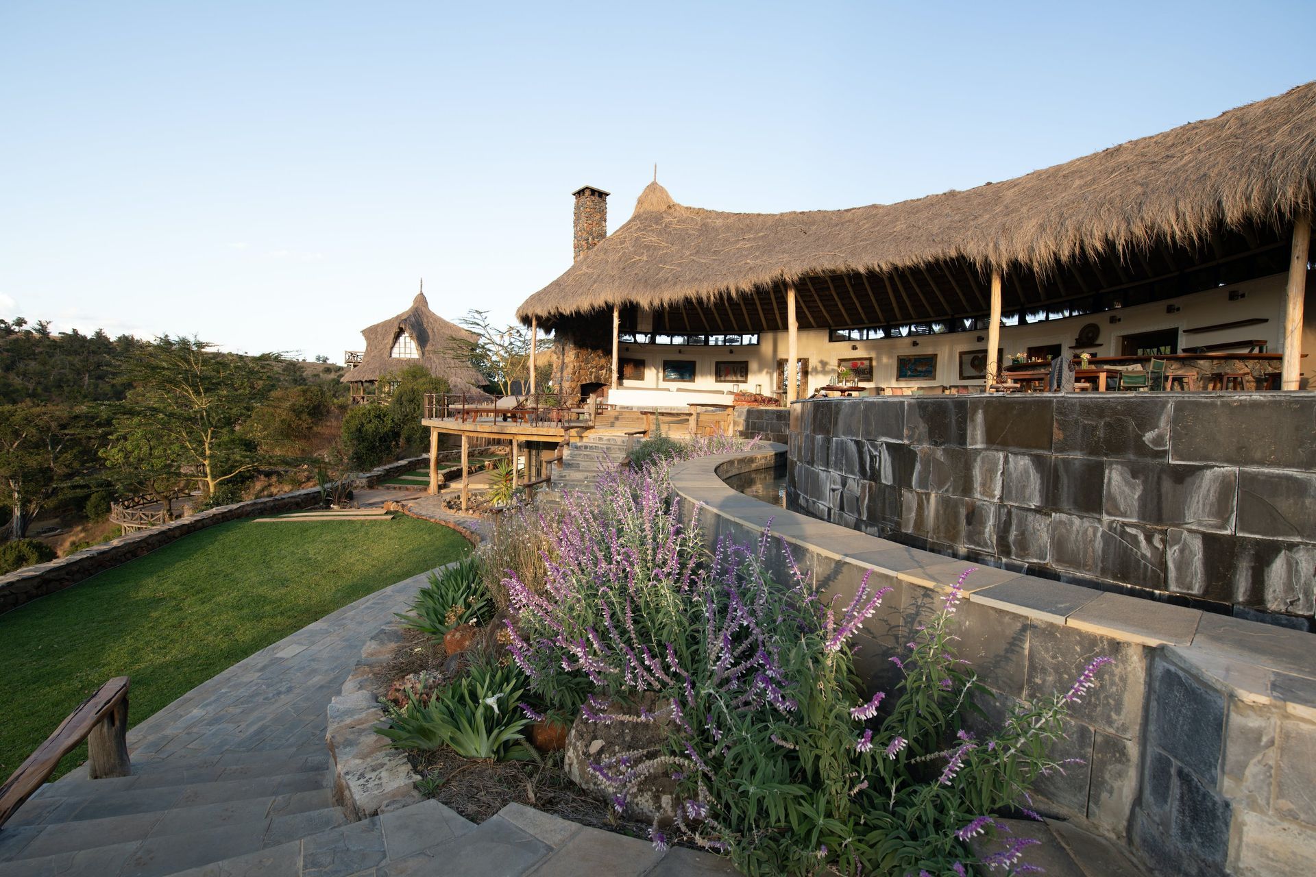 Thatched-roof building beside a flower-lined path and stone terrace at sunset