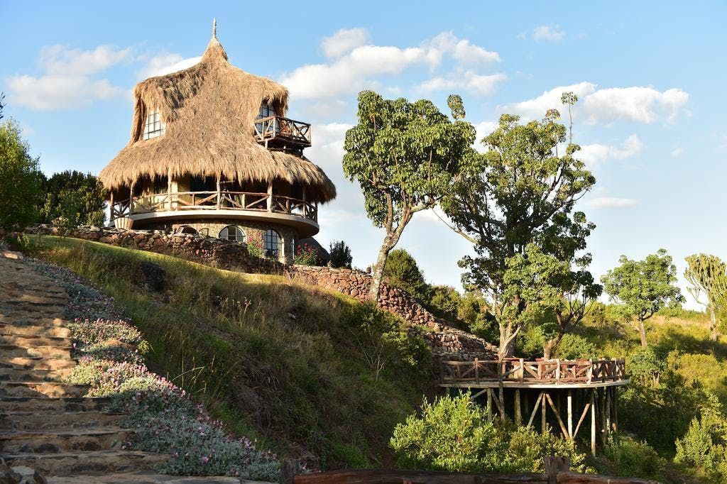 Thatched hut on a hillside with trees and a small wooden bridge under a blue sky