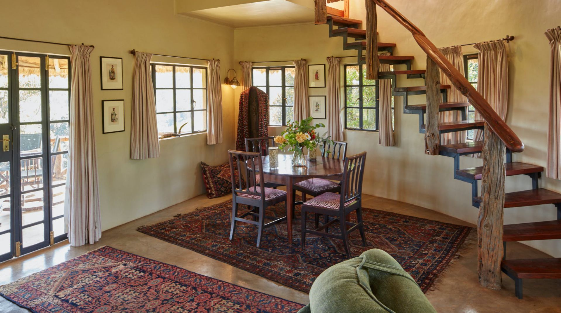 Cozy dining room with wooden table, patterned rugs, staircase, and French doors with sunlight.