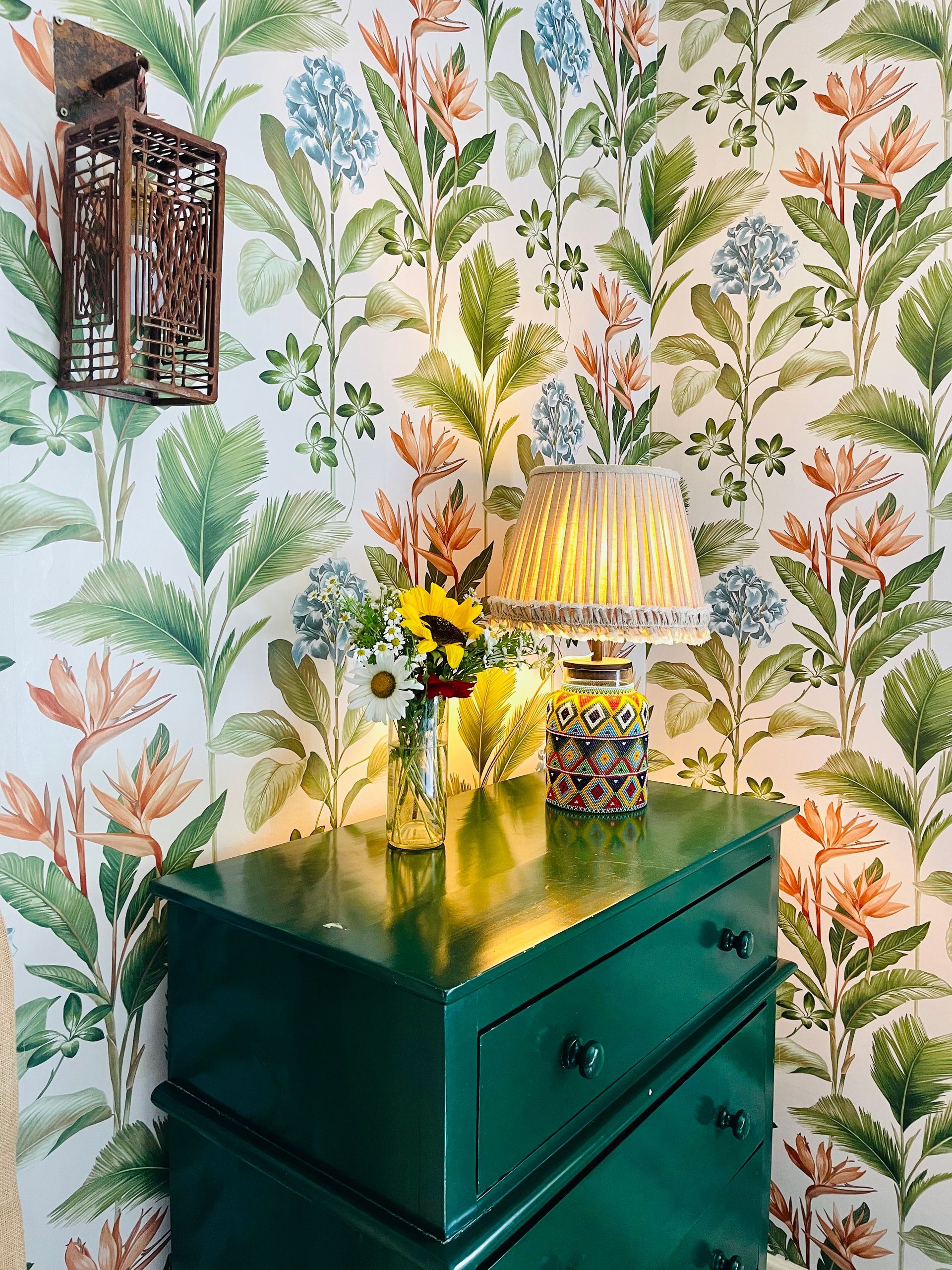 Green dresser with lamp and flowers against leafy patterned wallpaper