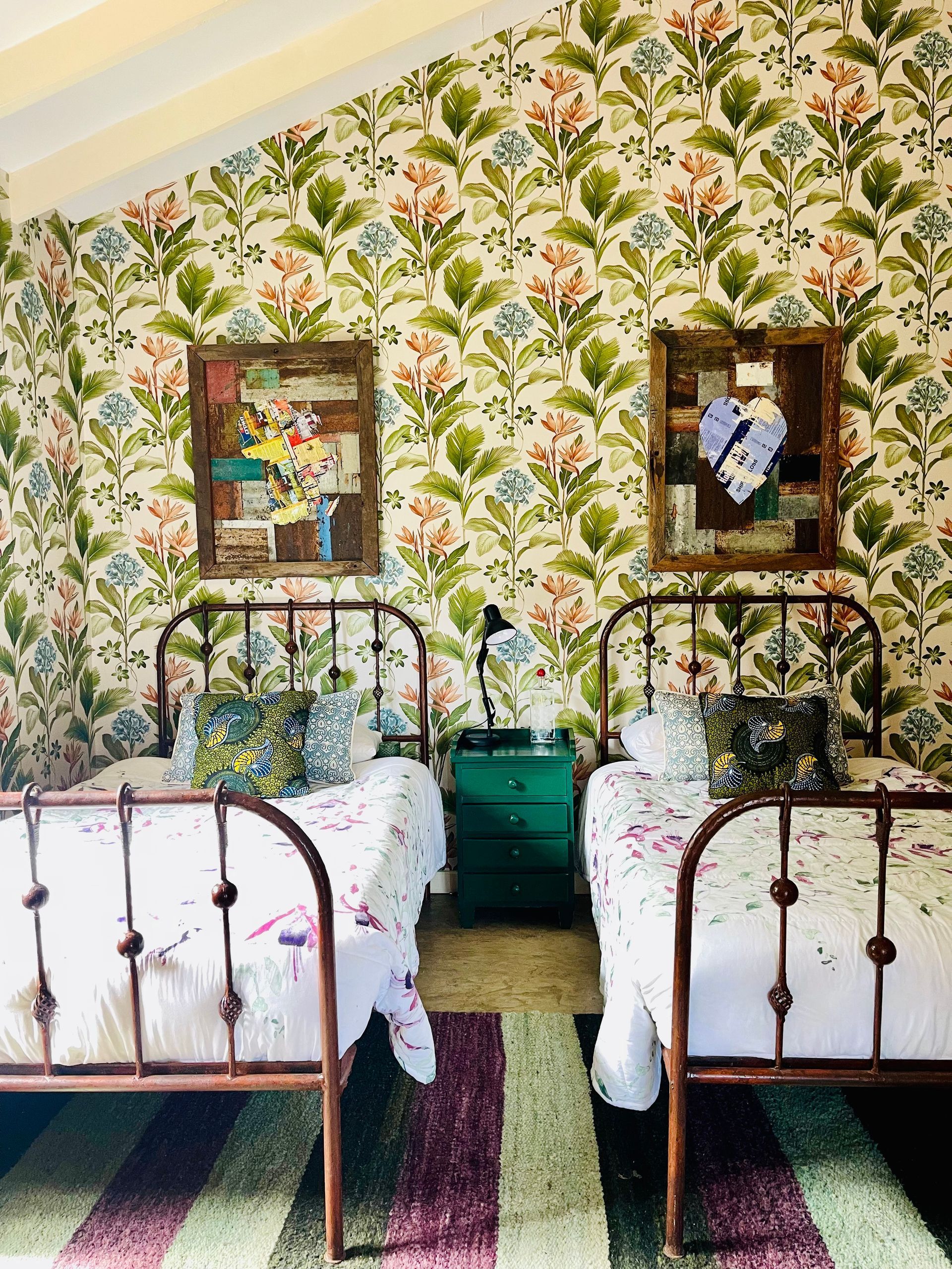 Two beds in a floral wallpapered room with striped rug, teal nightstand, and framed wall art