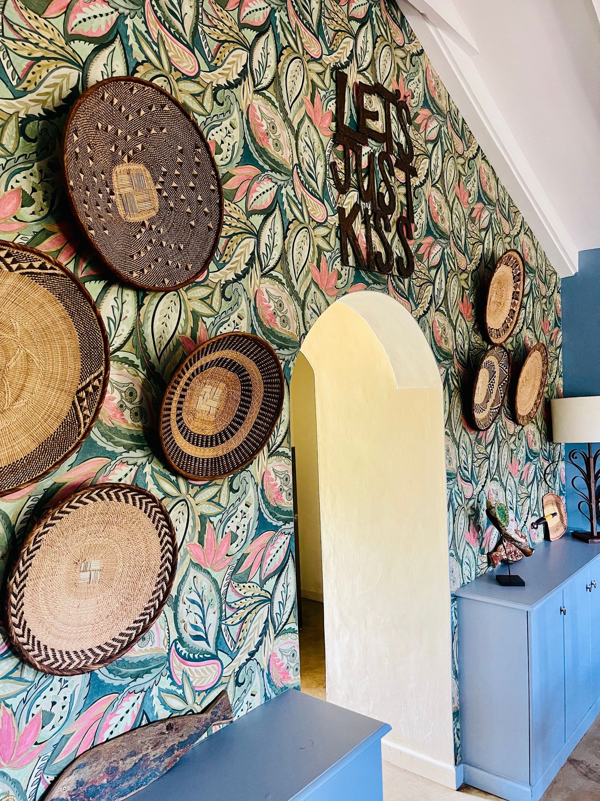 Colorful floral wallpaper with woven wall baskets, an arched doorway, and a blue cabinet in a hallway