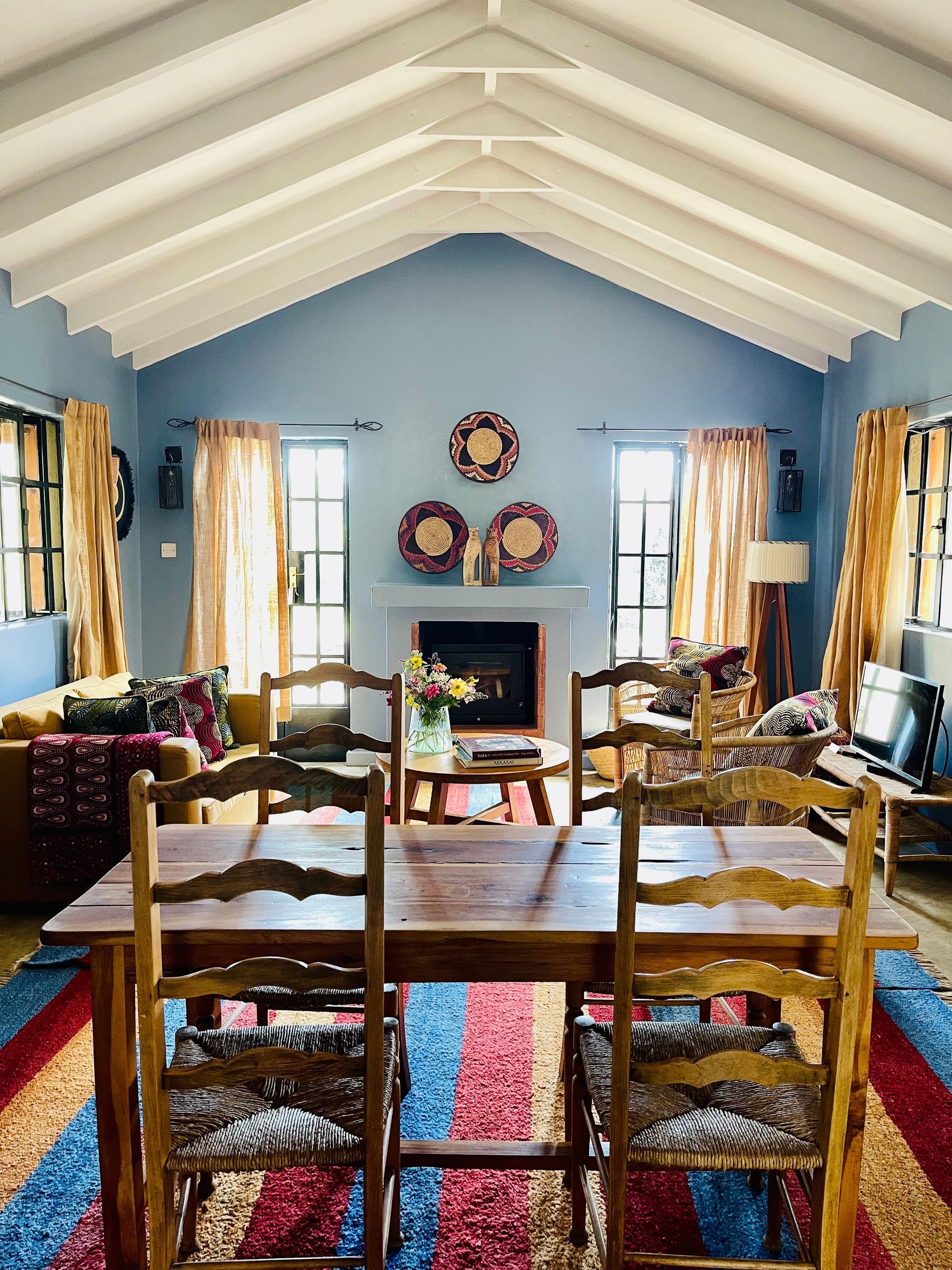 Cozy living room with blue walls, vaulted white ceiling, fireplace, and wooden dining table with chairs and colorful rugs