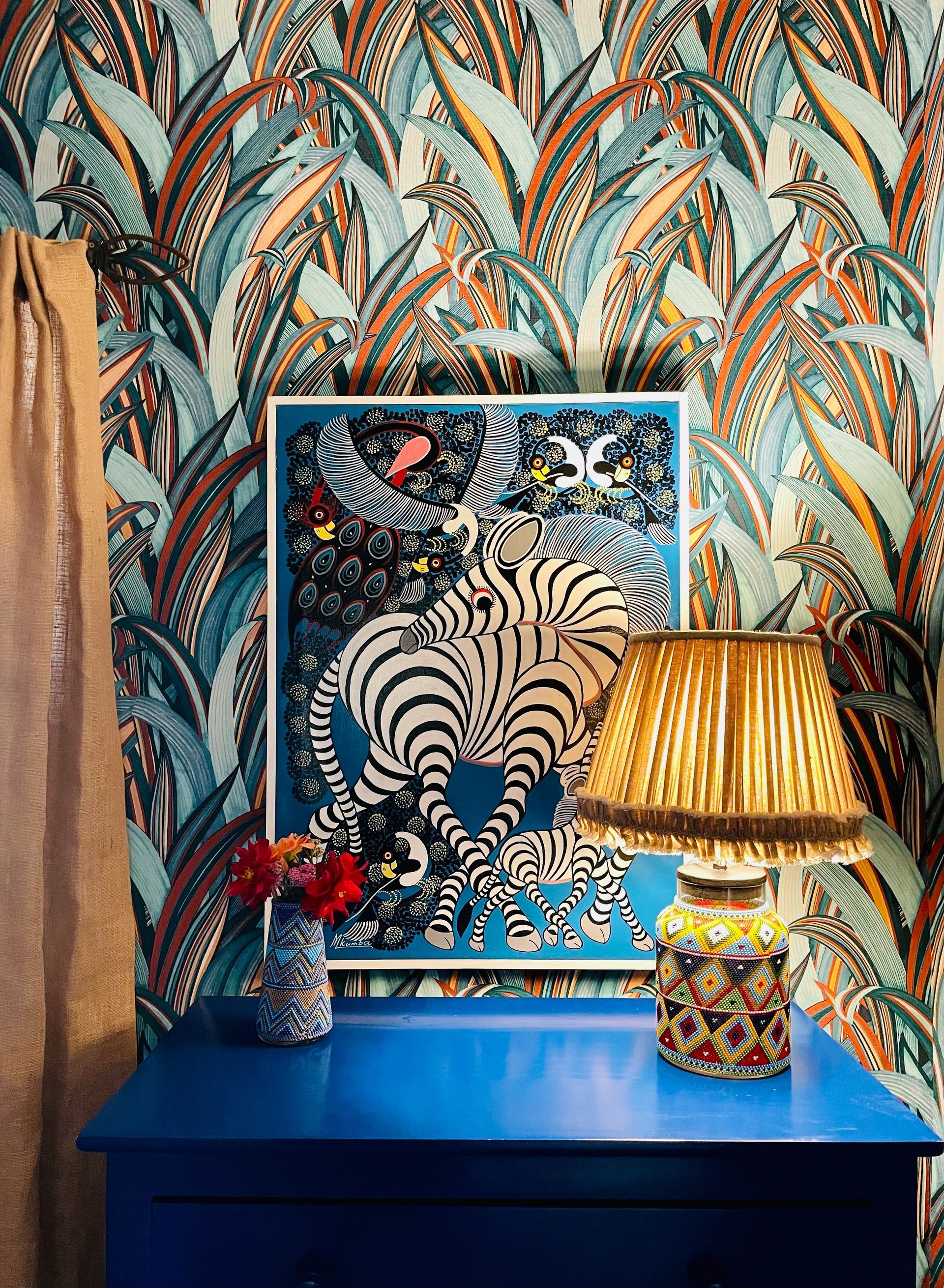 Blue console table with zebra art, tropical wallpaper, and a patterned lamp in a colorful room