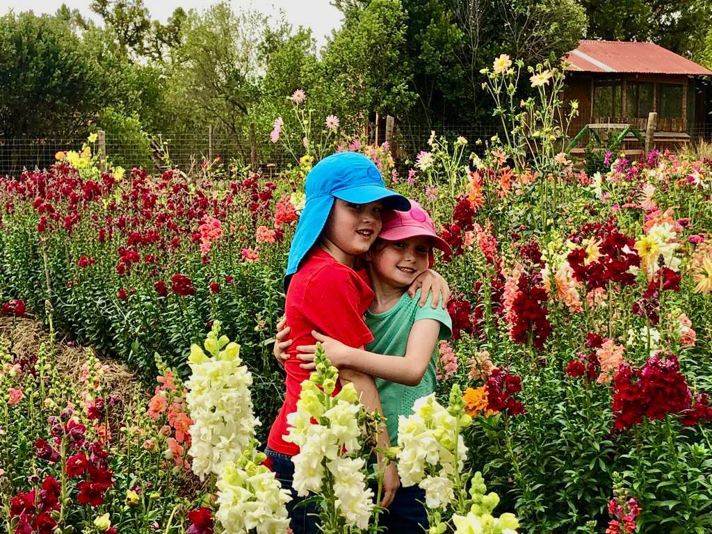 Two children hugging in a colorful flower garden, wearing blue and pink hats.
