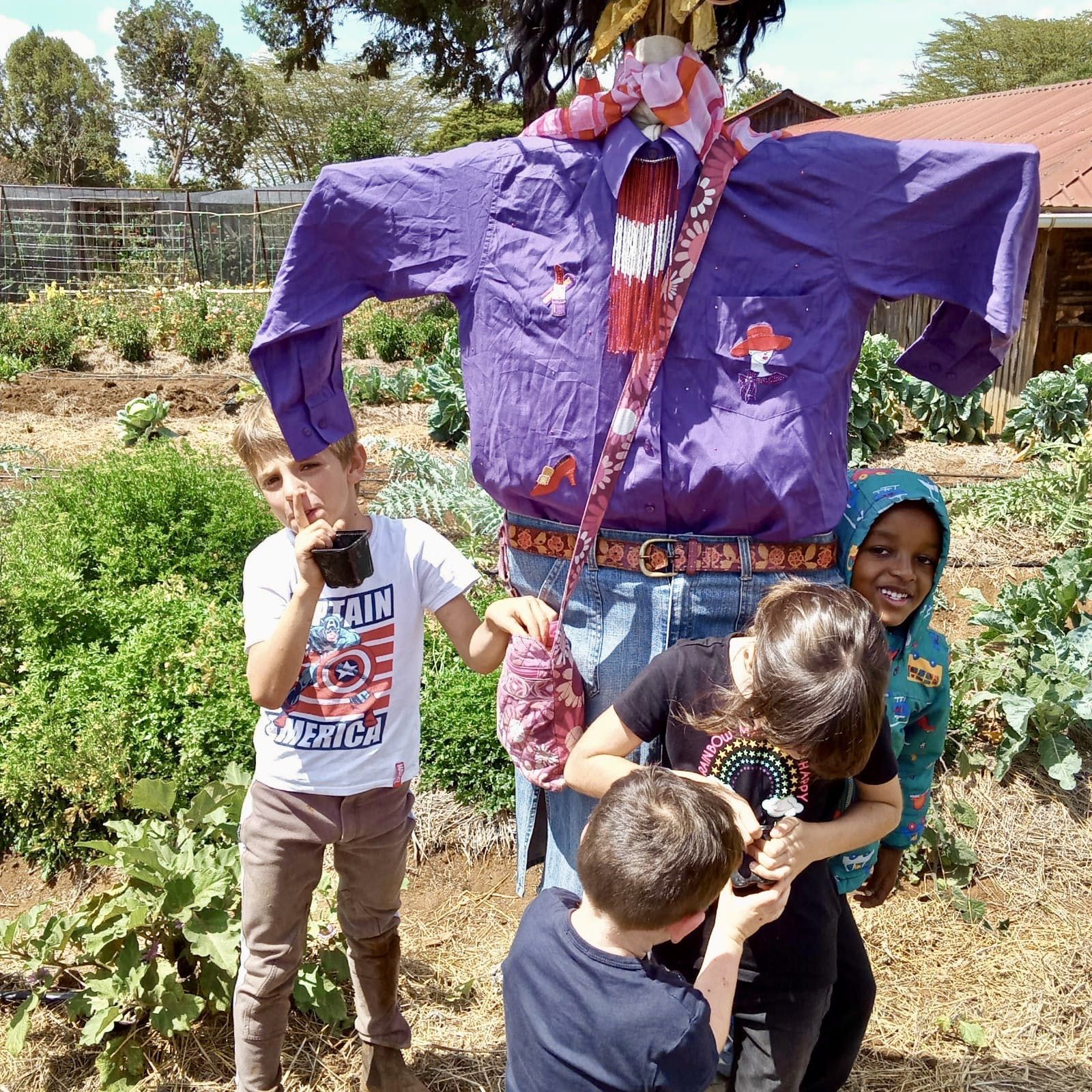 Children pose with a large purple scarecrow in a garden, one holding a smaller figure.