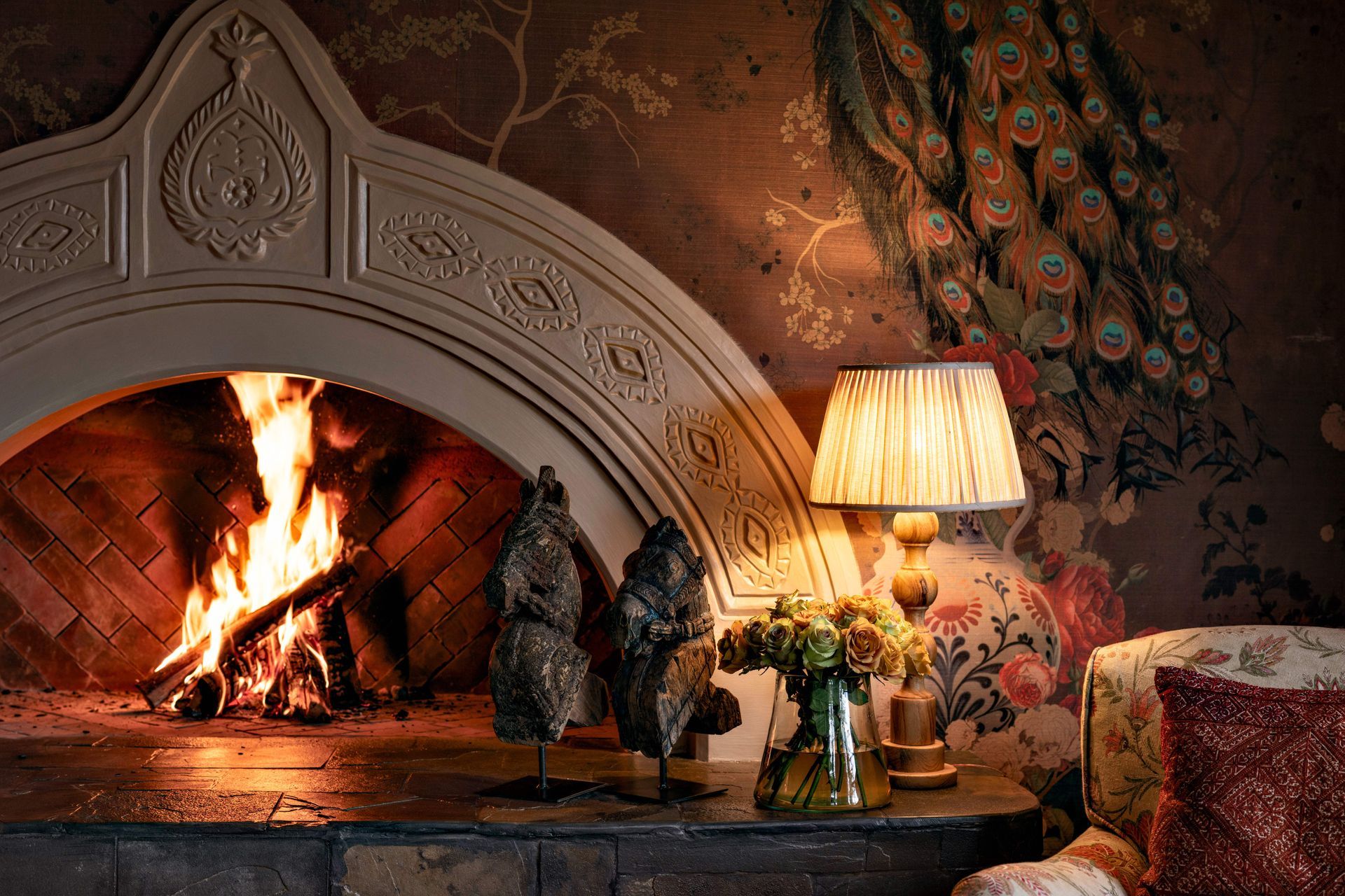 Cozy fireplace with a bright fire, ornate mantel, lamp, and flowers in a warmly lit room
