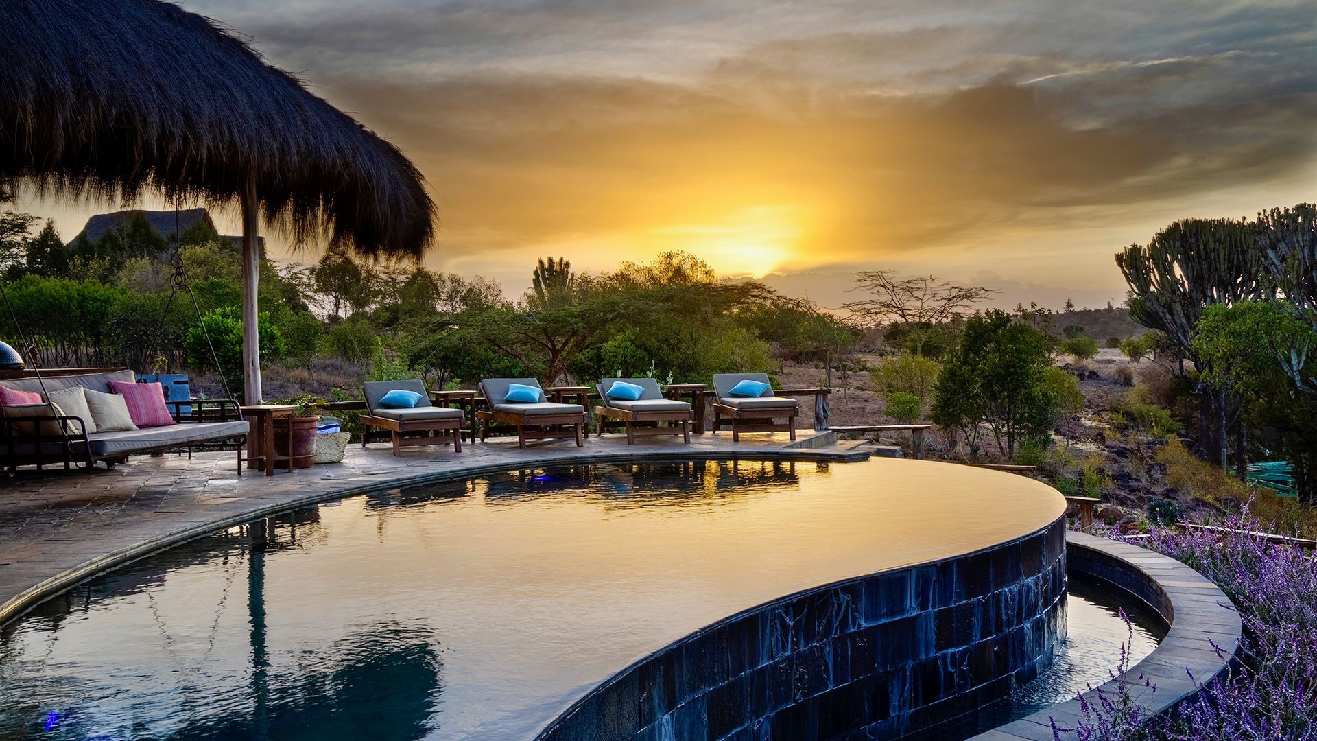 Sunset over a resort pool with lounge chairs, palm thatch umbrellas, and a tropical hillside view