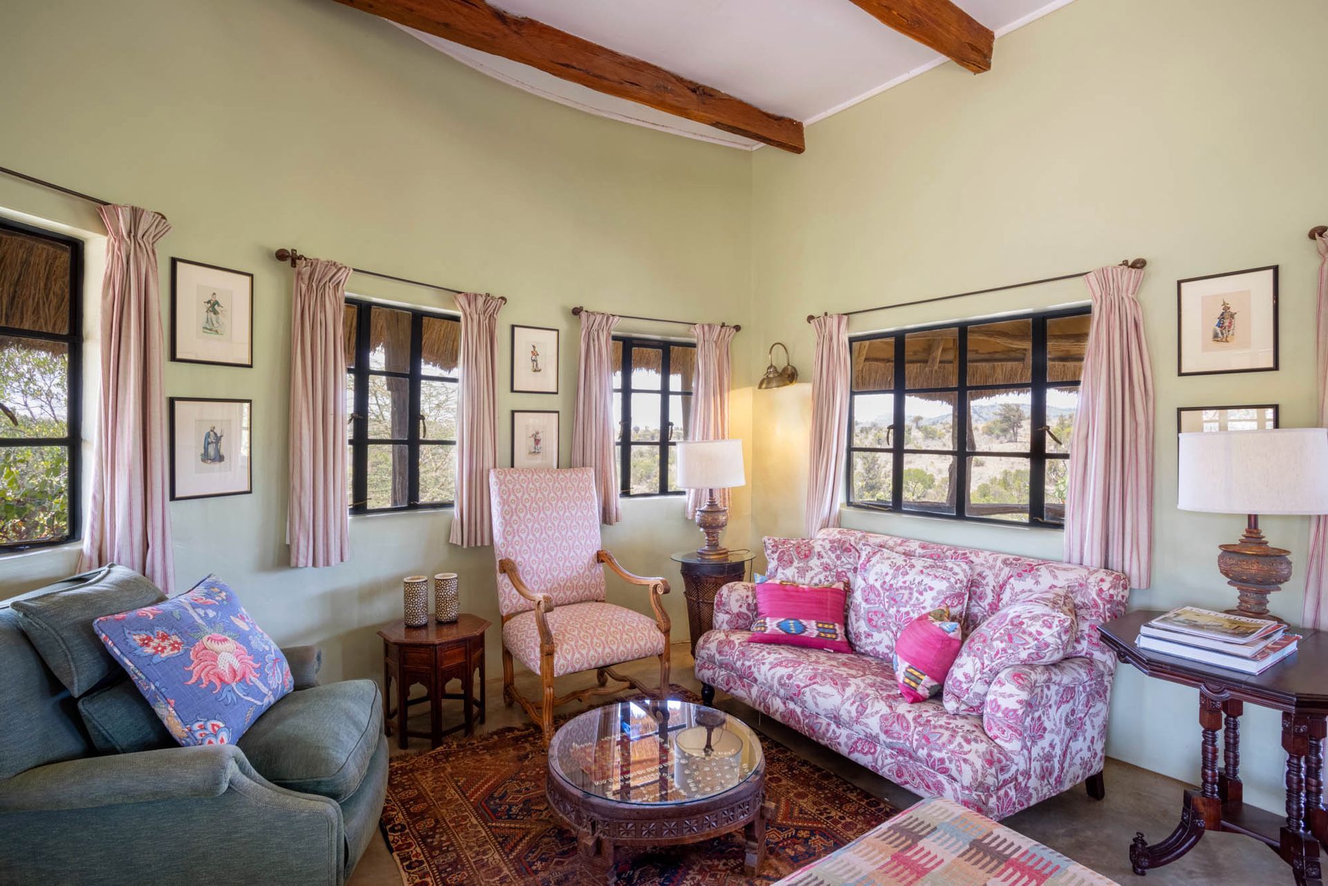 Cozy living room with floral sofa, green armchair, wood beams, and large windows with patterned curtains