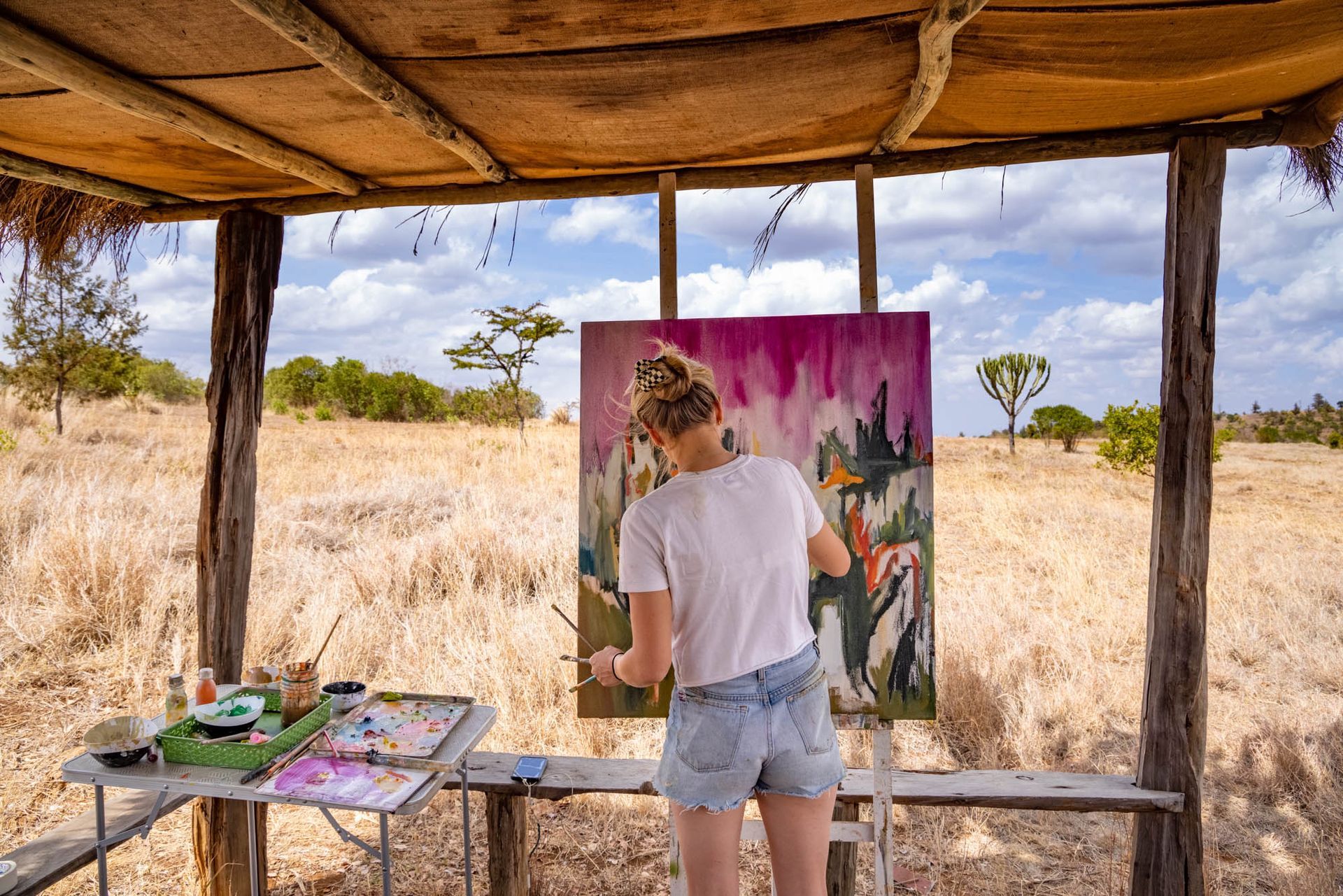 Person painting at an easel in a rustic open-air studio overlooking dry grassland.