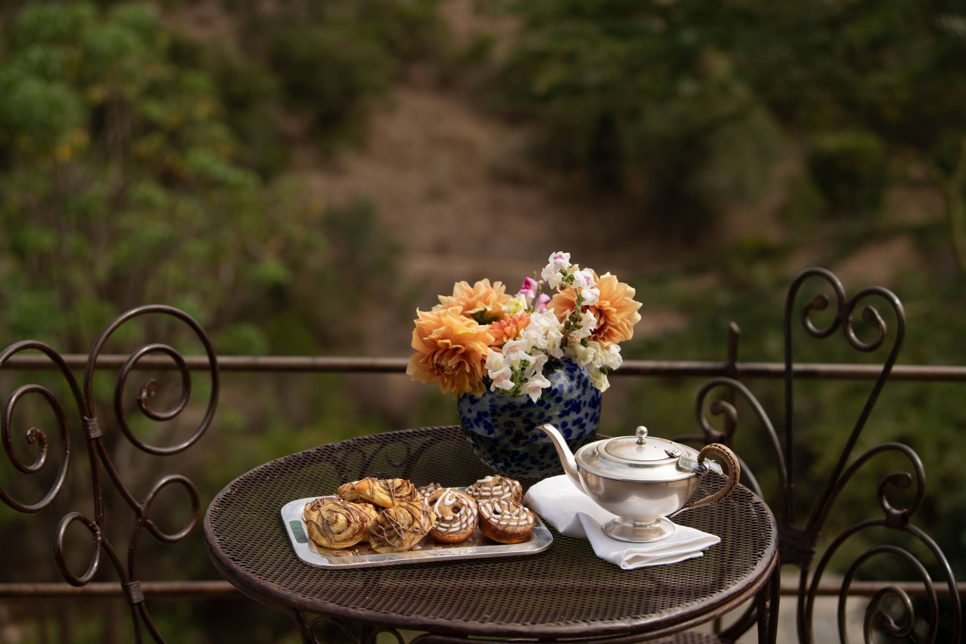 Outdoor patio table with tea set, pastries, and a vase of orange flowers against a blurred garden backdrop