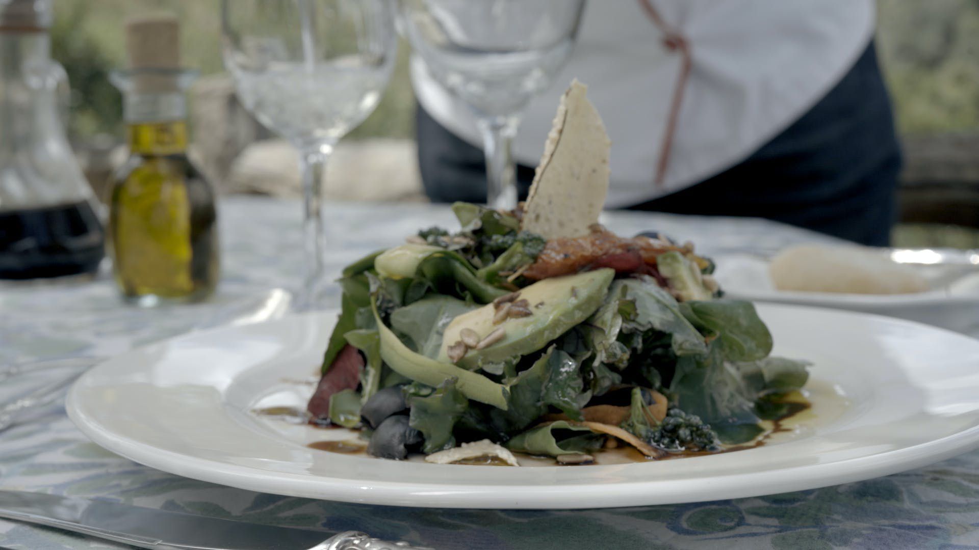 Plate of mixed salad with greens and cheese on a dining table, with wine glasses and olive oil in the background