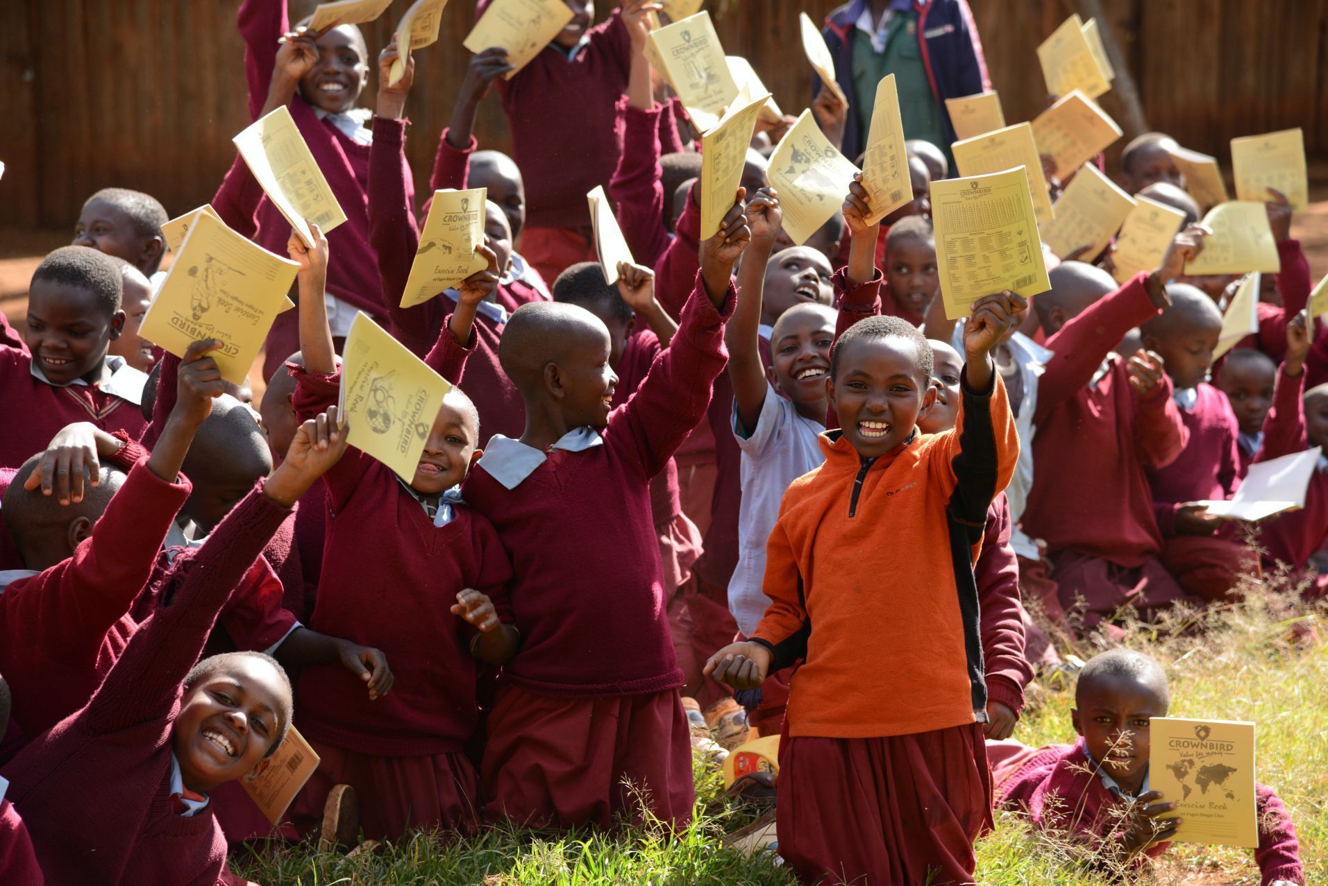 Children in maroon uniforms holding yellow papers outdoors, one in an orange shirt smiling in front