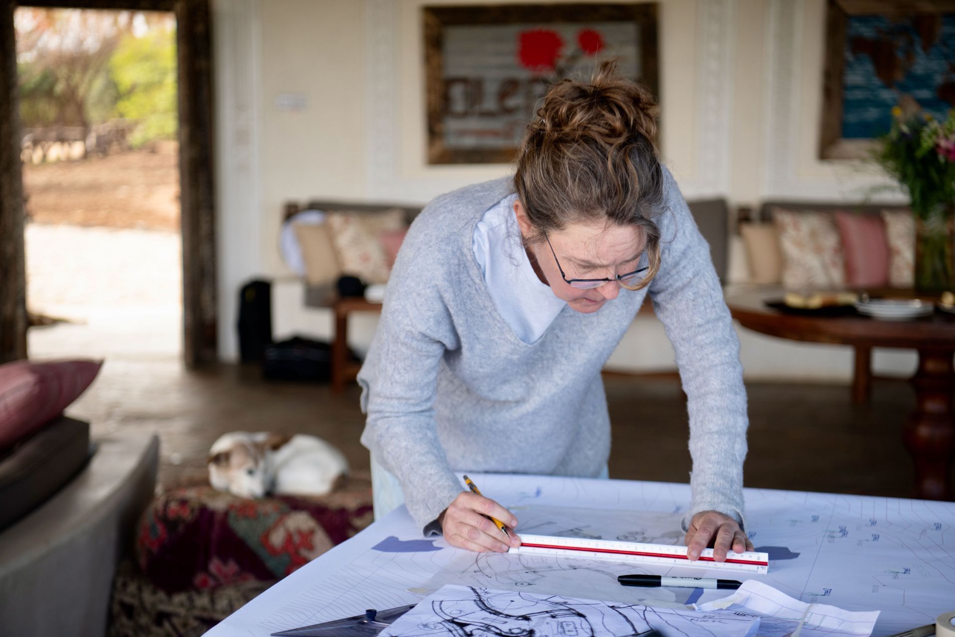 Person leaning over a table, writing on papers in a bright living room.