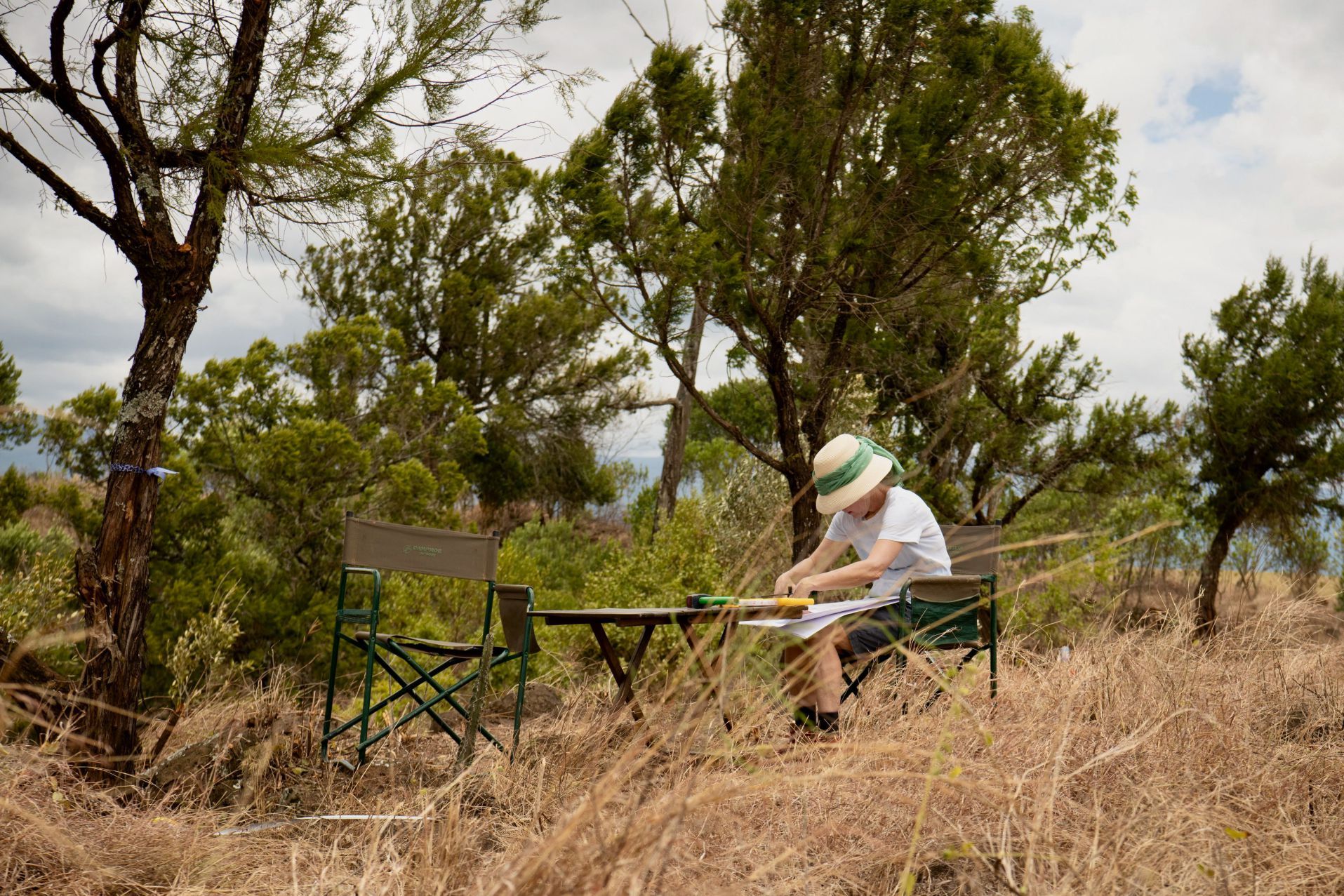 Person sitting at a folding table in a dry wooded field, wearing a hat and writing or drawing.