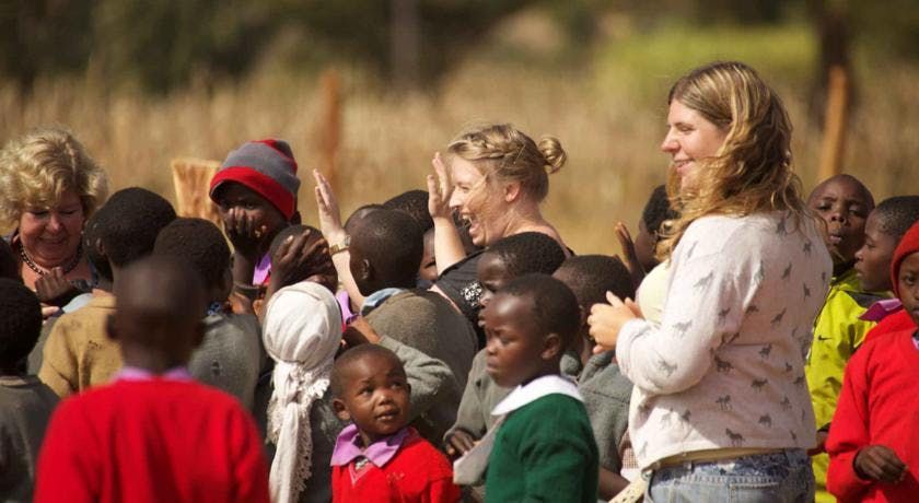 Group of children and two adults gathered outdoors in daylight, including one woman in a white sweater.