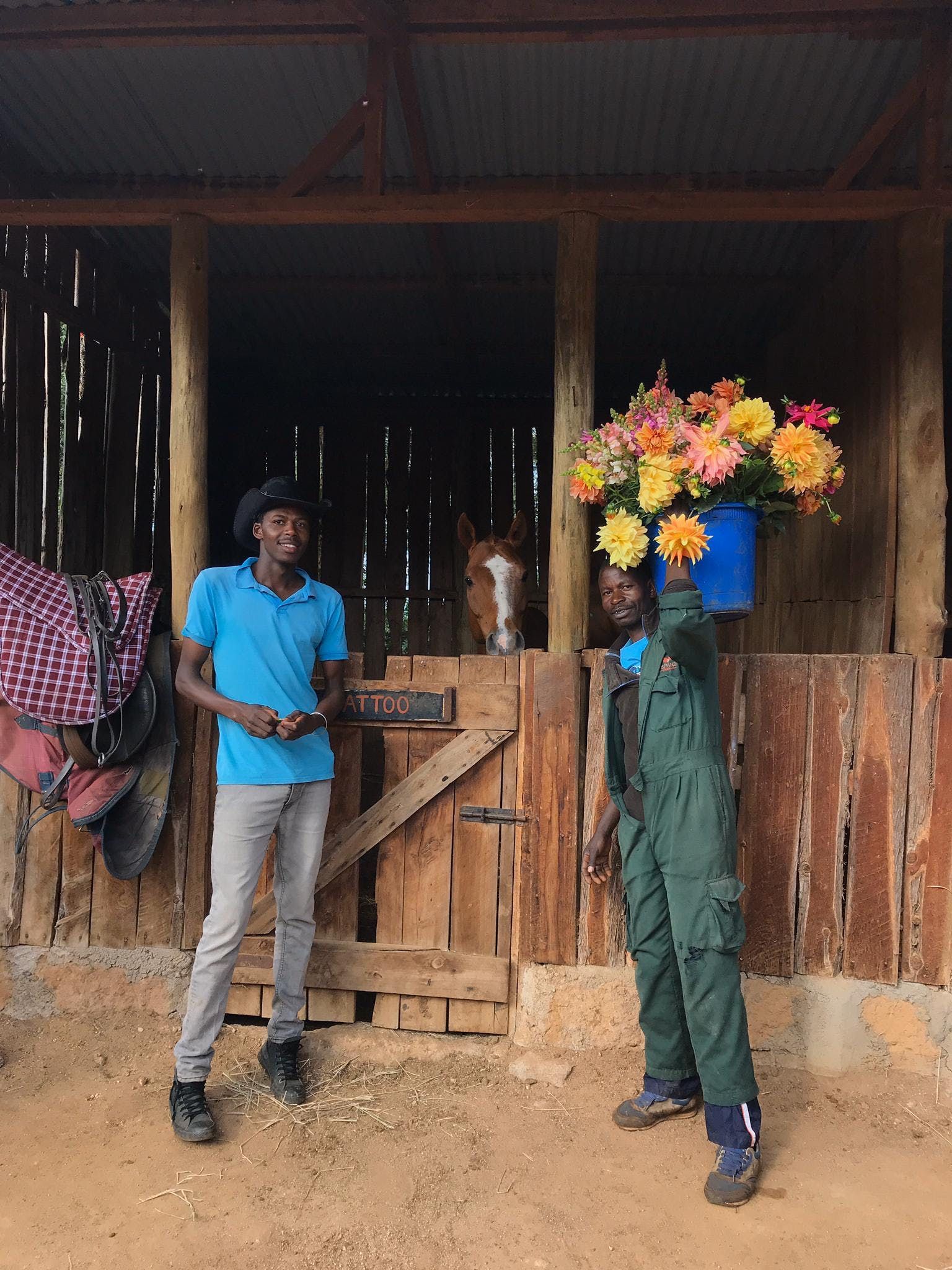 Two people stand by a wooden shed; one has a bouquet of colorful flowers covering their head.
