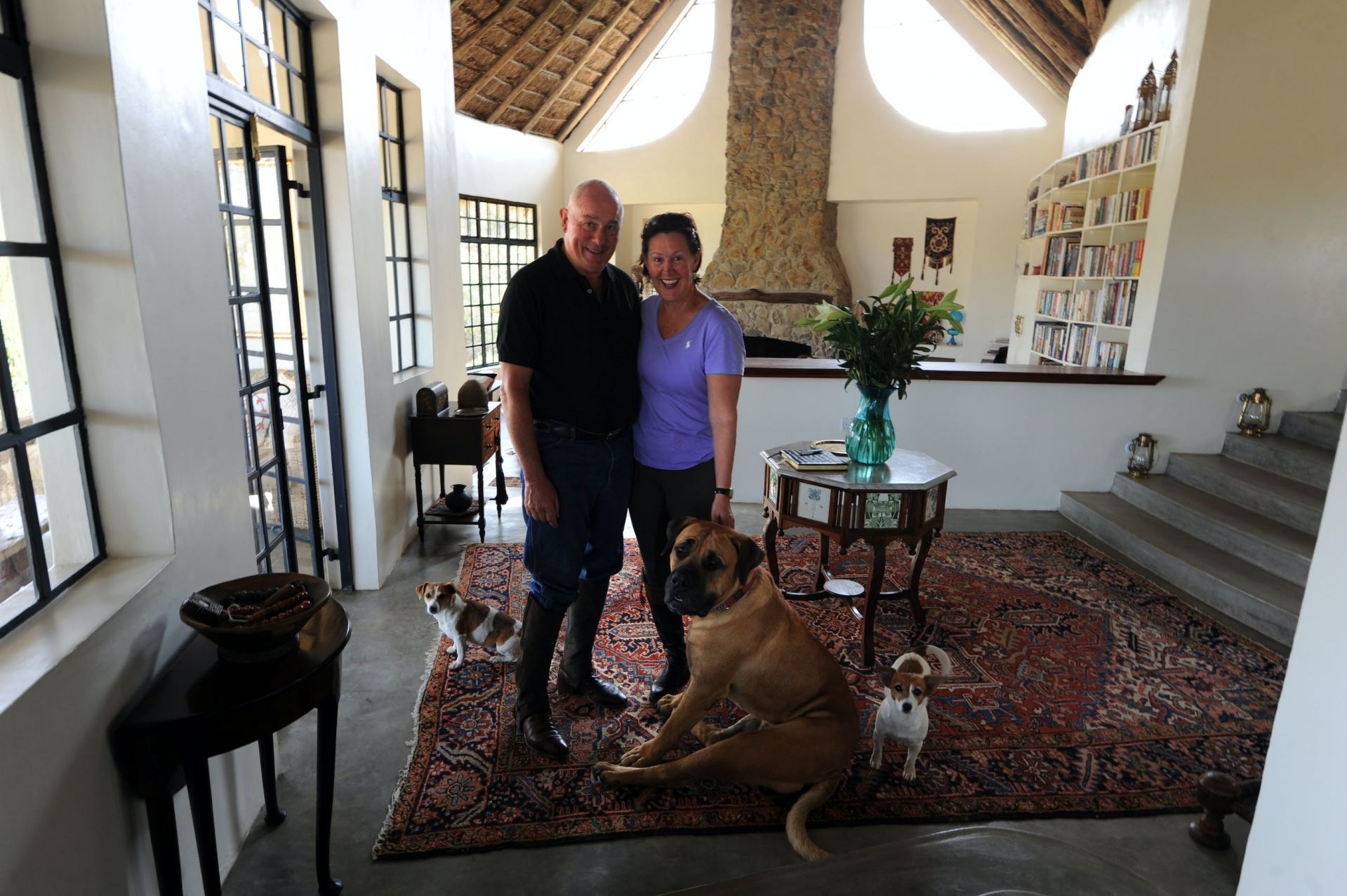 Couple standing with a large dog in a sunlit living room with patterned rug and stairs