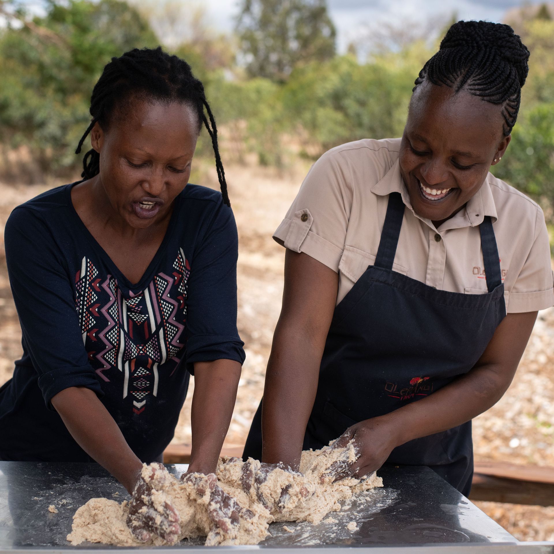 Two people knead dough together at an outdoor table, smiling in a bright setting.