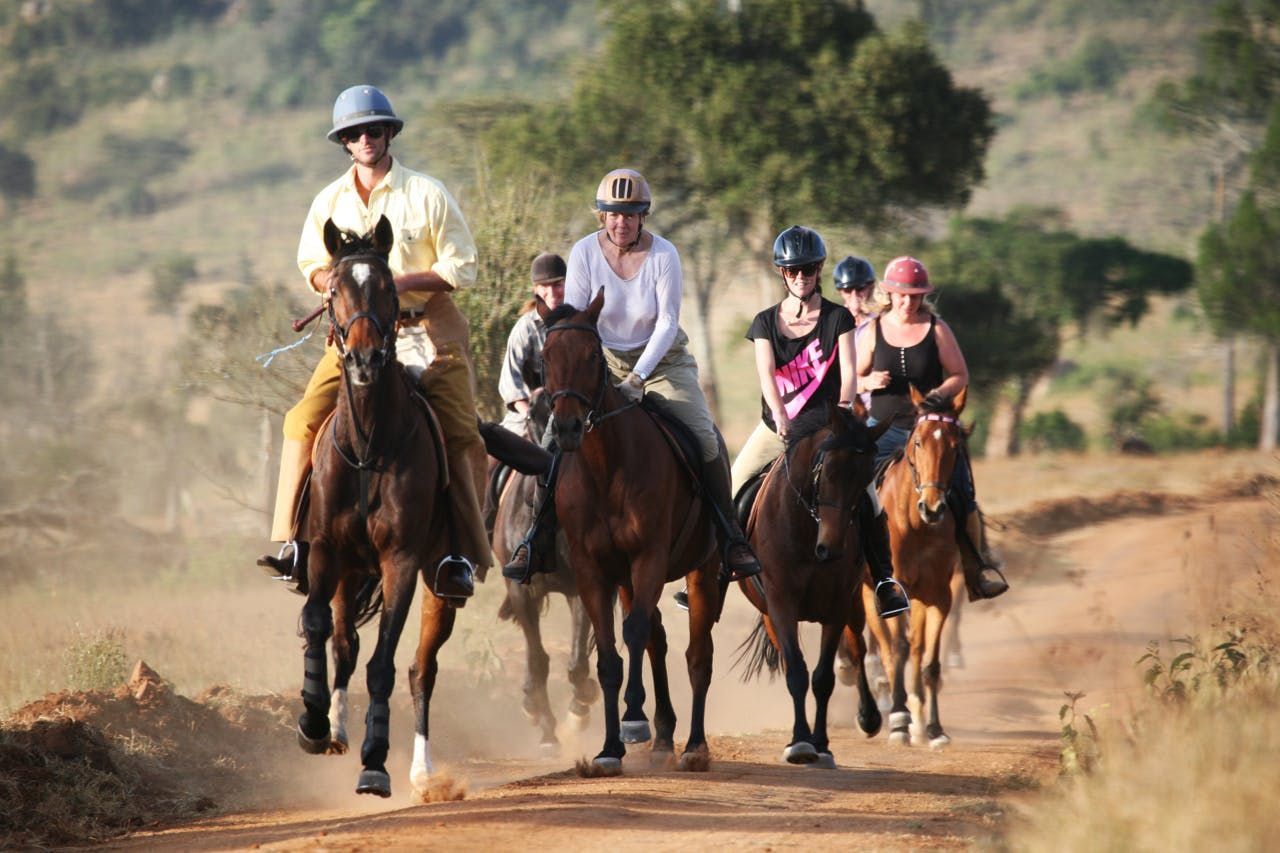Group of riders on horseback kicking up dust along a dirt trail in a dry, hilly landscape.