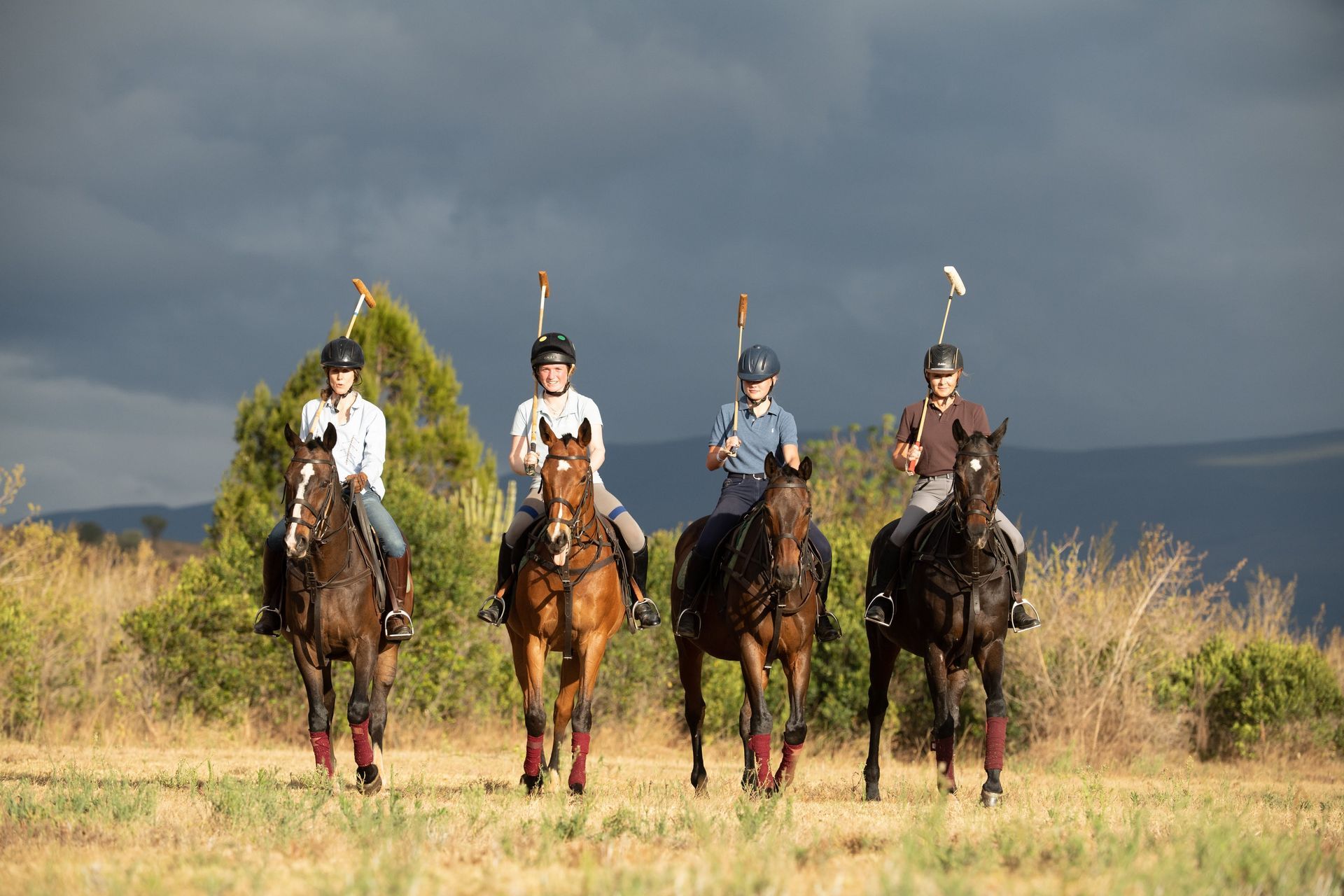 Four riders on horseback in a grassy field under a dark, cloudy sky.