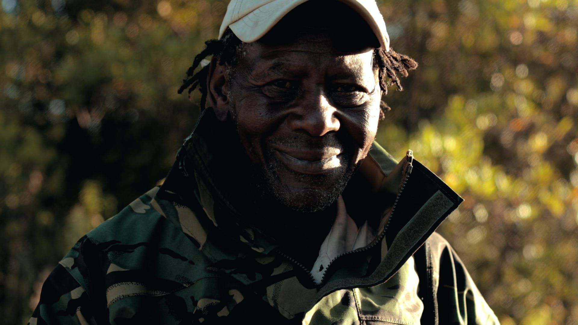 Smiling man in a camouflage jacket and cap standing outdoors in sunlight.