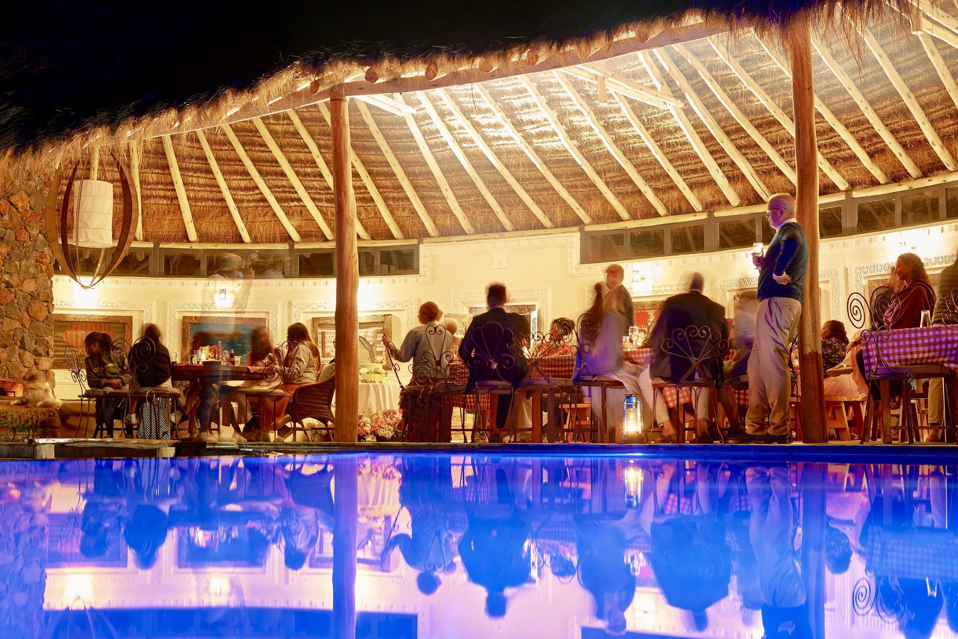 Open-air restaurant at night with diners under warm lights reflected in a blue-lit pool