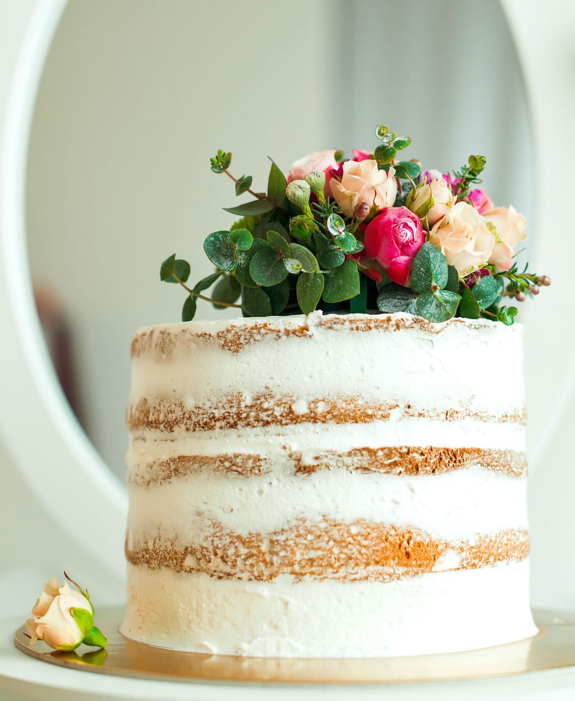 Rustic white frosted cake topped with pink flowers and greenery on a light table
