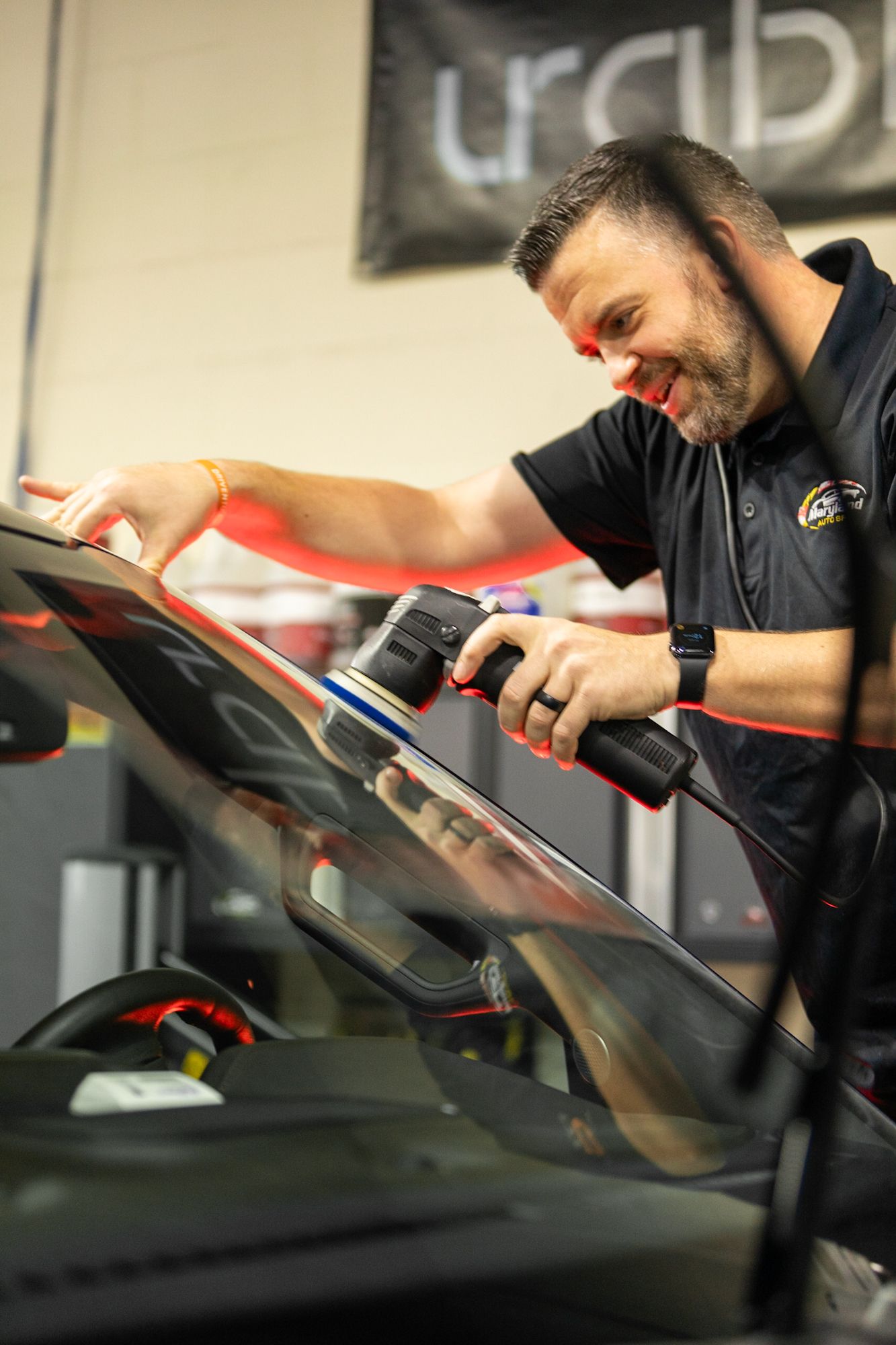 A smiling technician uses a power buffer to polish the windshield of a vehicle in an auto shop.