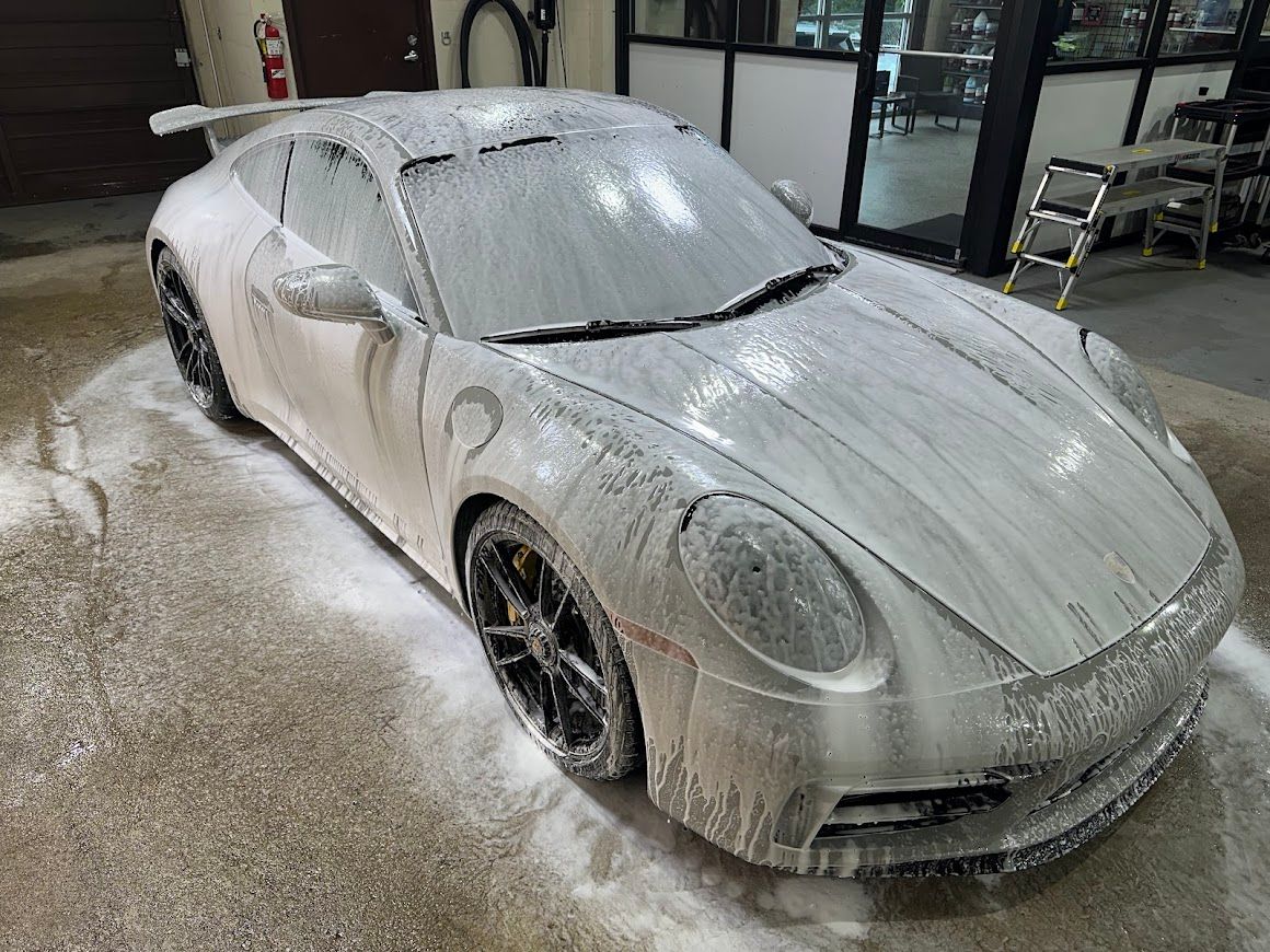 A high-angle view of a white Porsche sports car covered in thick, white soap suds inside a professional auto detail shop.