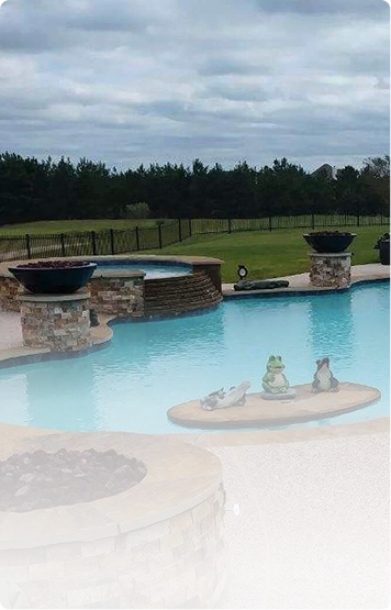 Backyard pool with stone fire pits, grassy lawn, and cloudy sky beyond the water.