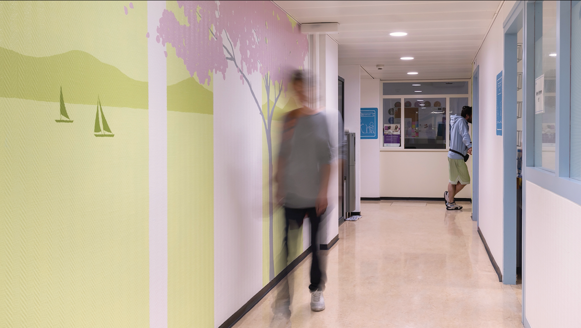 Blurred person walking in a bright hallway with pale walls and doors lining both sides.