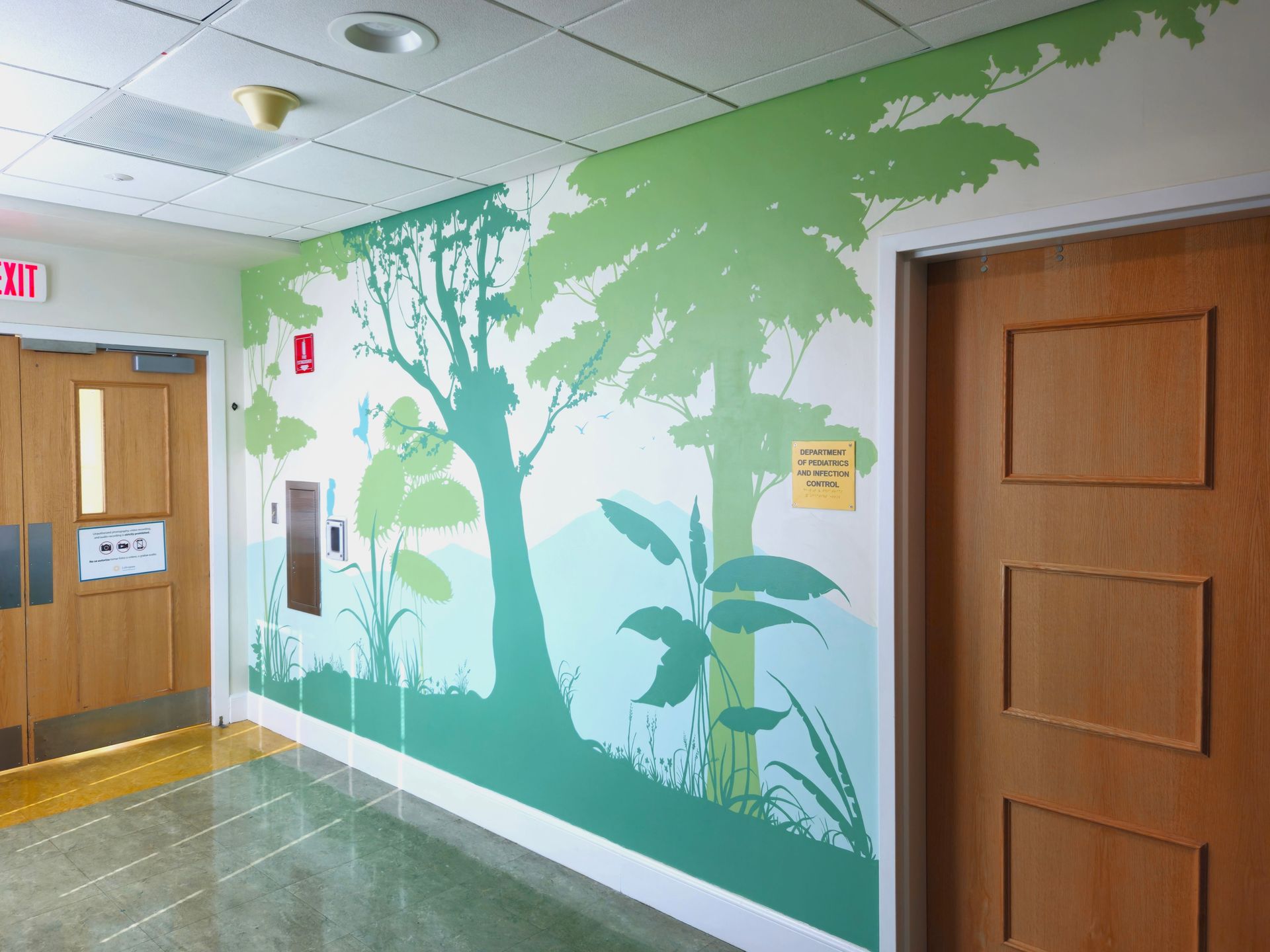Hallway wall mural with green trees and leaves beside wooden doors and exit sign