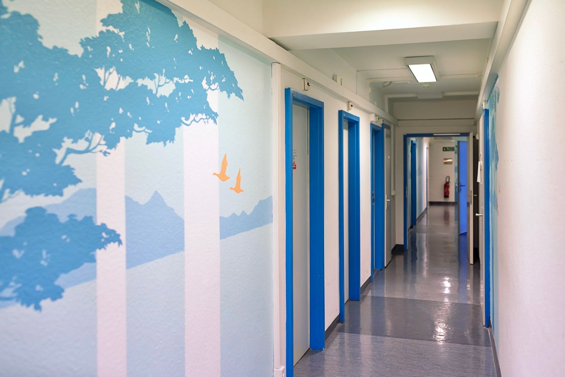Bright hospital hallway with blue doors and a sky-and-cloud mural on the left wall