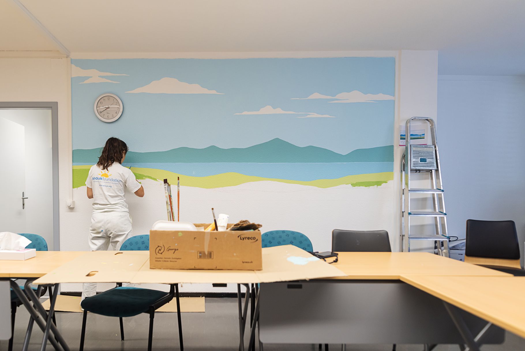 Person painting a mountain mural in a bright meeting room with tables, chairs, and a ladder