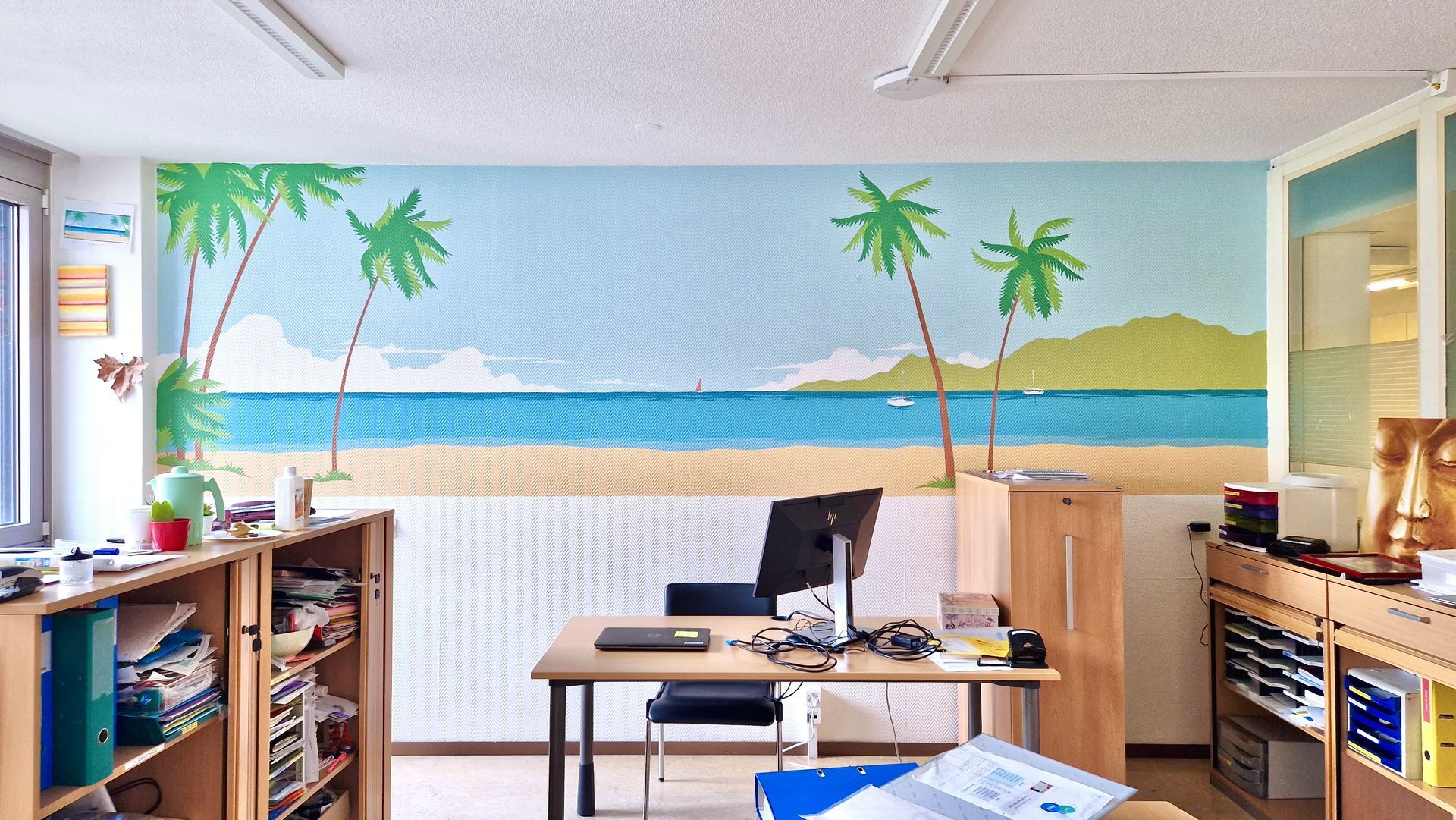 Office with a beach mural, desks, shelves, and computer equipment inside a bright room.
