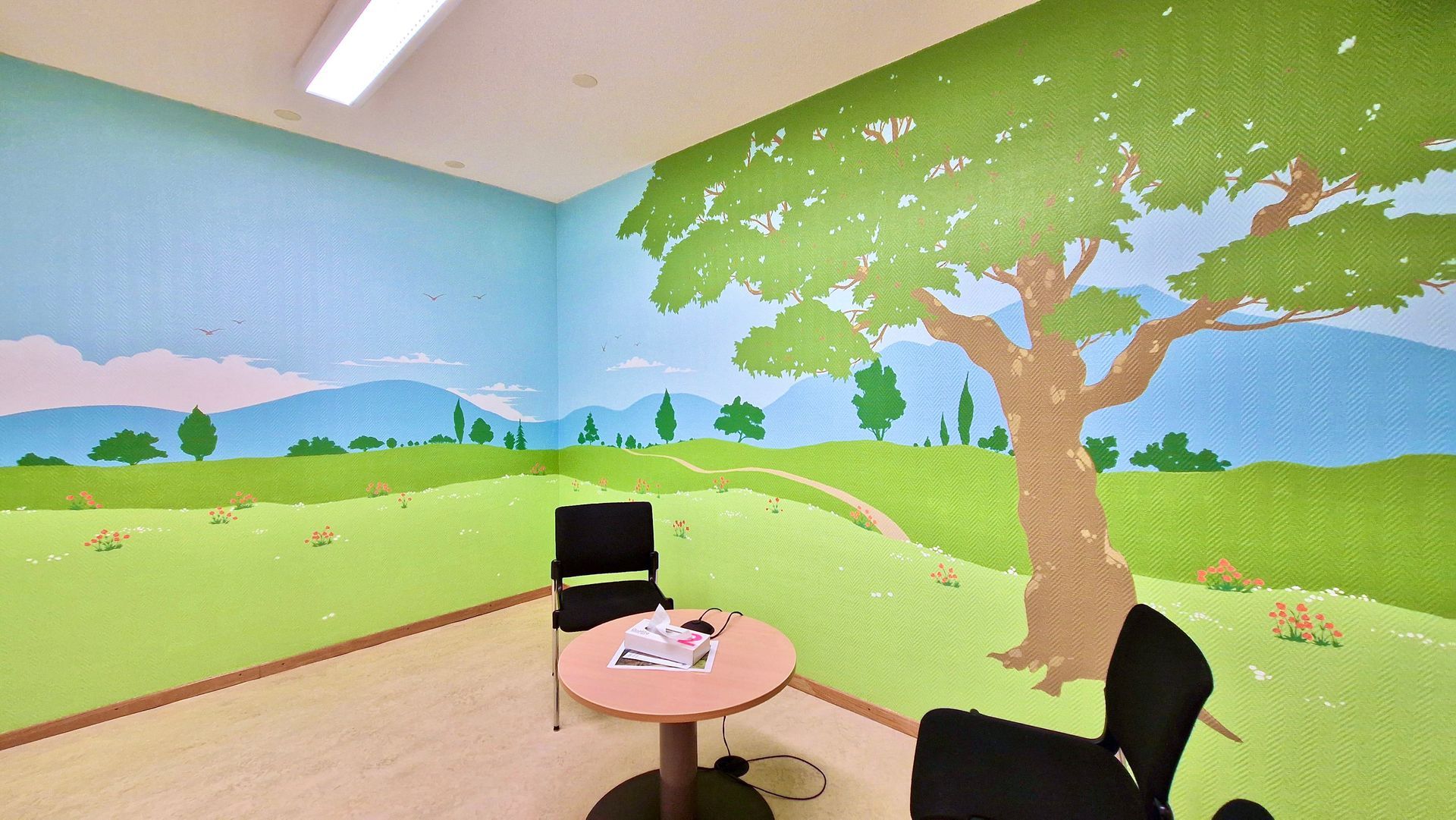 Mural-covered room with a tree and green landscape, featuring two black chairs and a small round table.