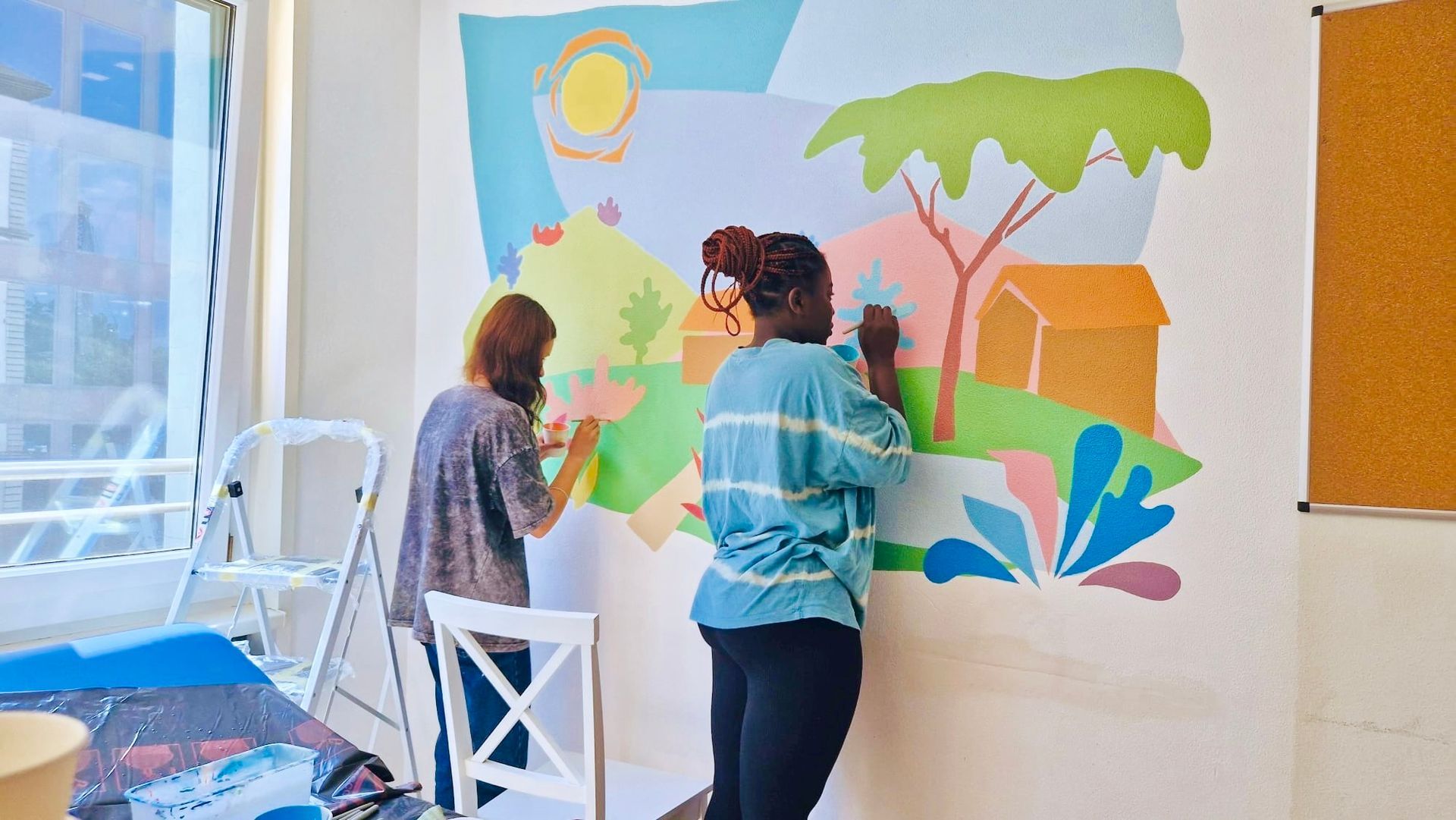 Two people painting a colorful mural on a wall in a bright room