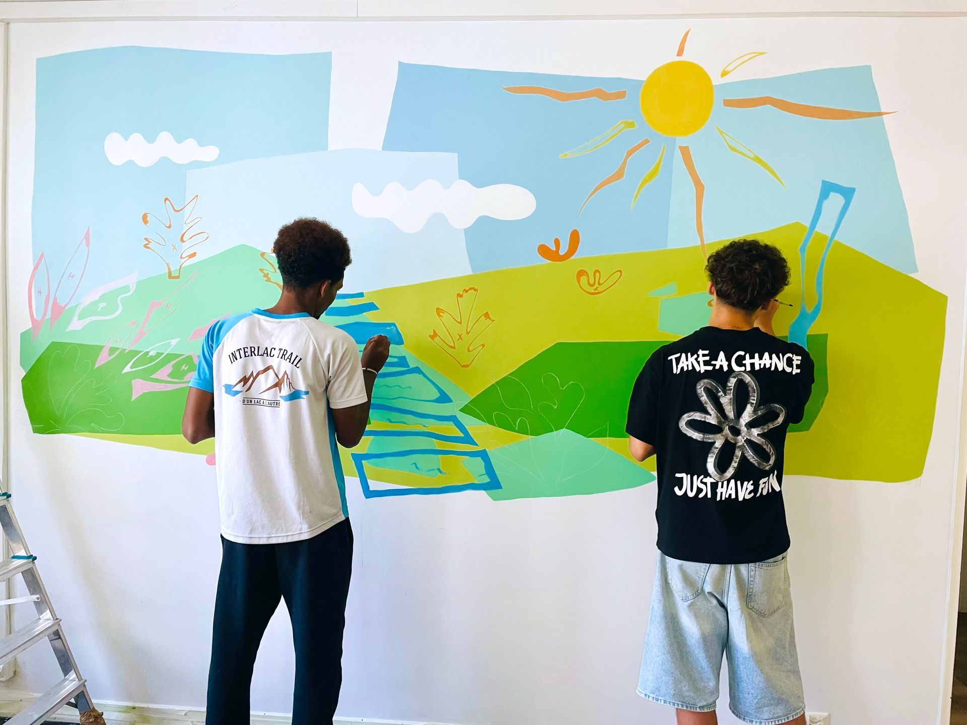 Two people painting a colorful mural with sun, clouds, grass, and a blue stream on a wall