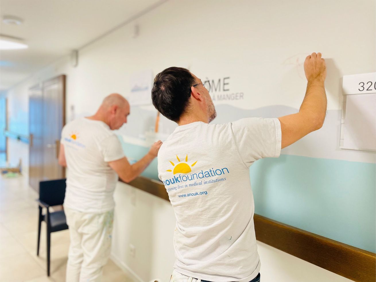 Two workers hang a sign in a bright hallway, standing beside a white wall and handrail.